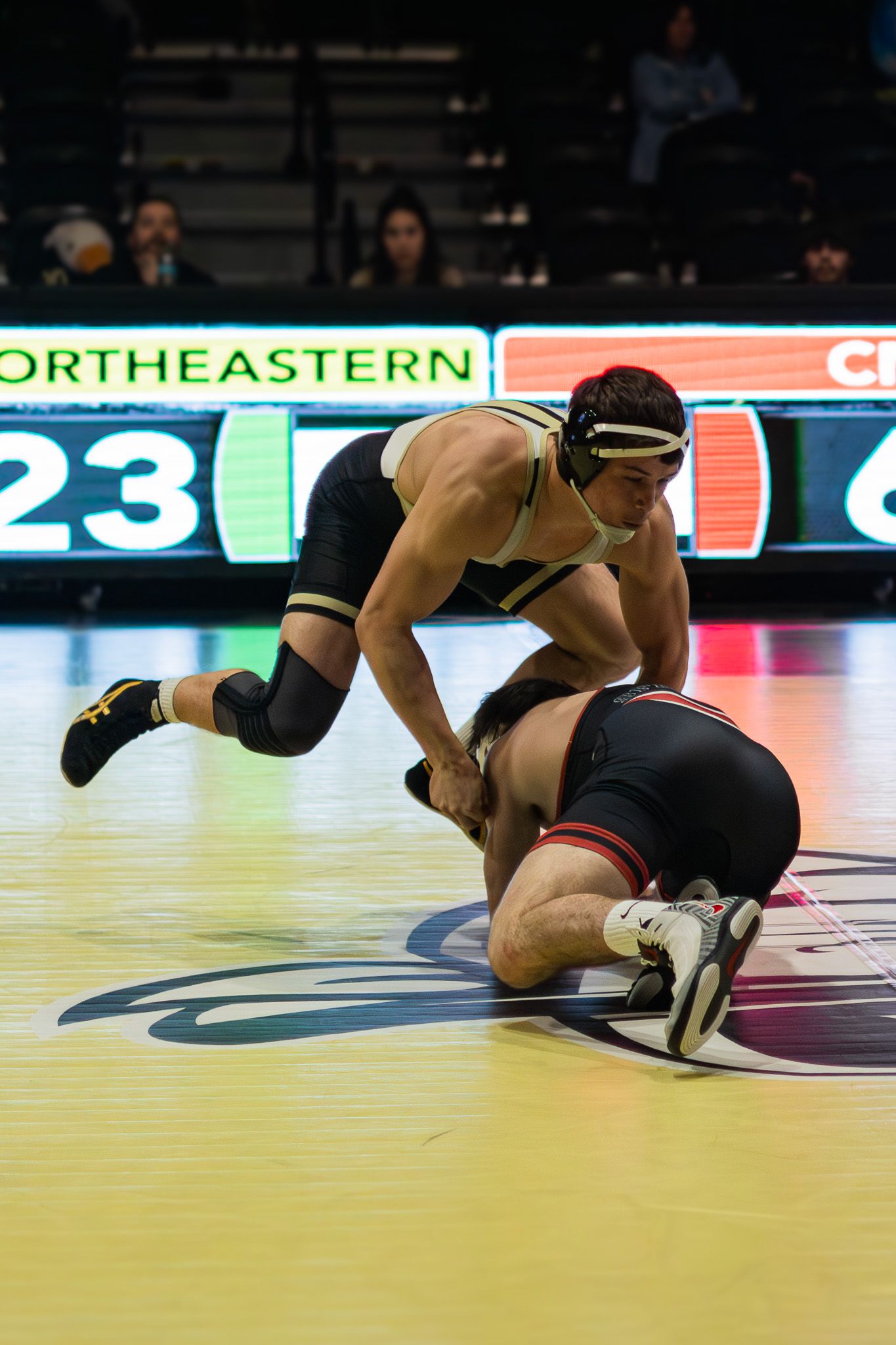 Wrestler in gold singlet takes down opponent on mat at Northeastern university wrestling match.