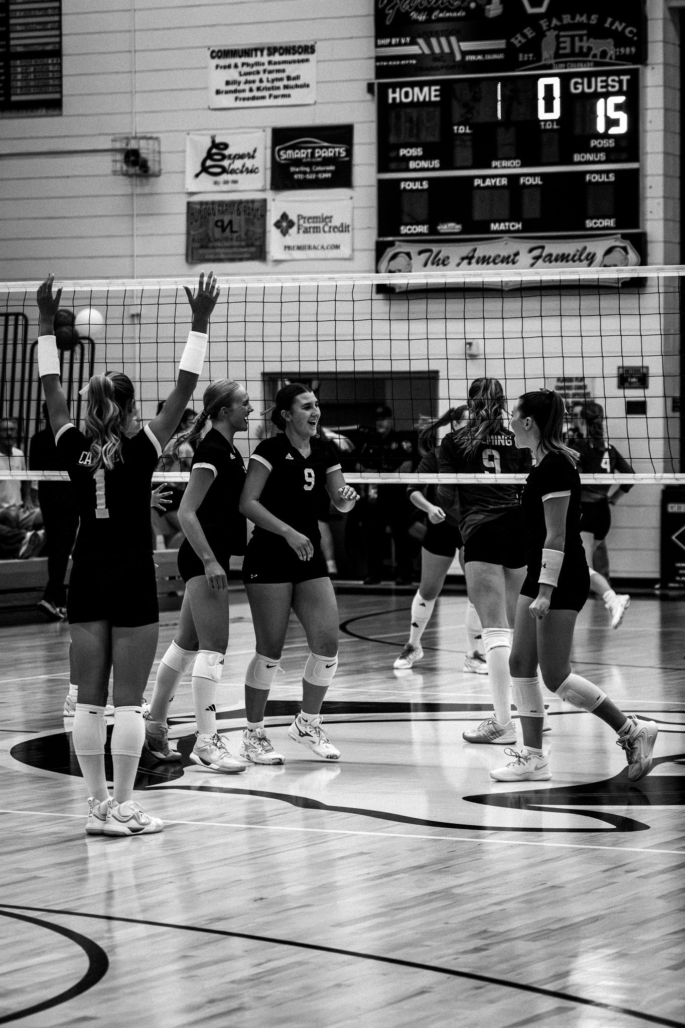 Female volleyball team in black uniforms celebrating during a match with scoreboard visible in background.