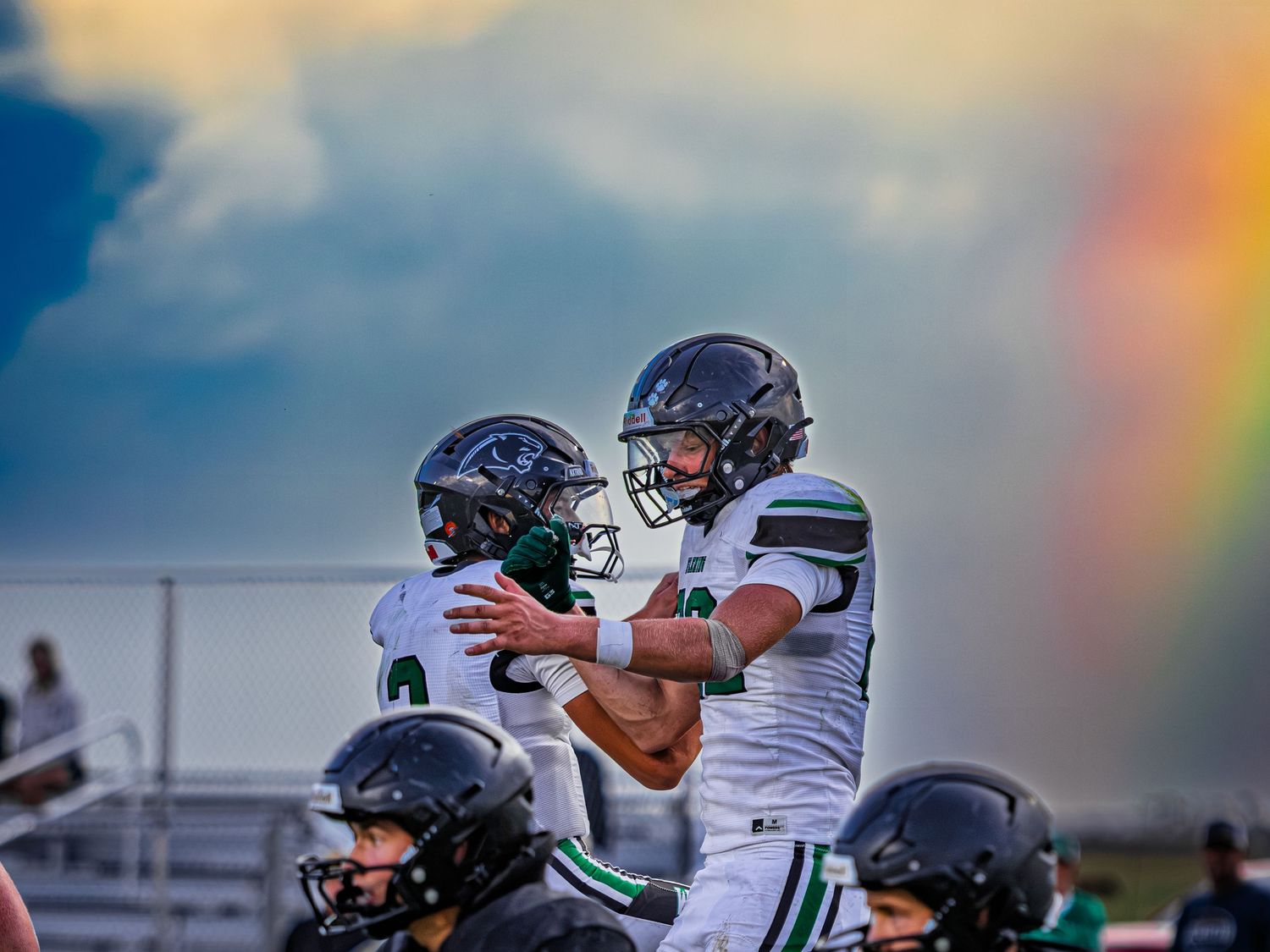 Two football players in green and white uniforms celebrate together with a colorful rainbow visible in background.