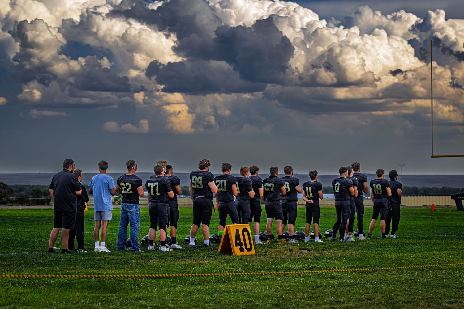 Football team lined up on green field at dusk with dramatic storm clouds filling the sky behind them.