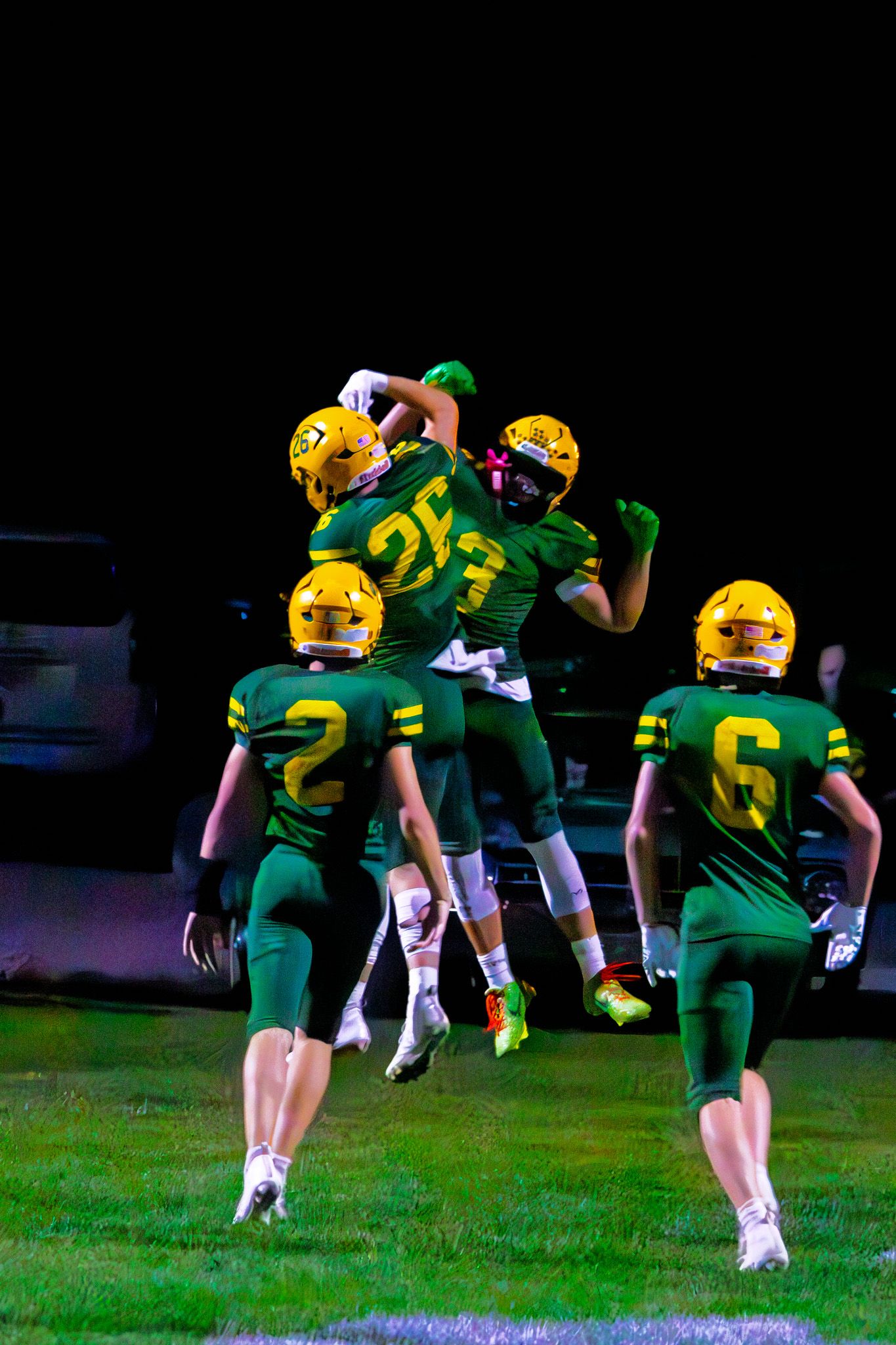 Green and gold high school football players celebrating a touchdown under Friday night lights.