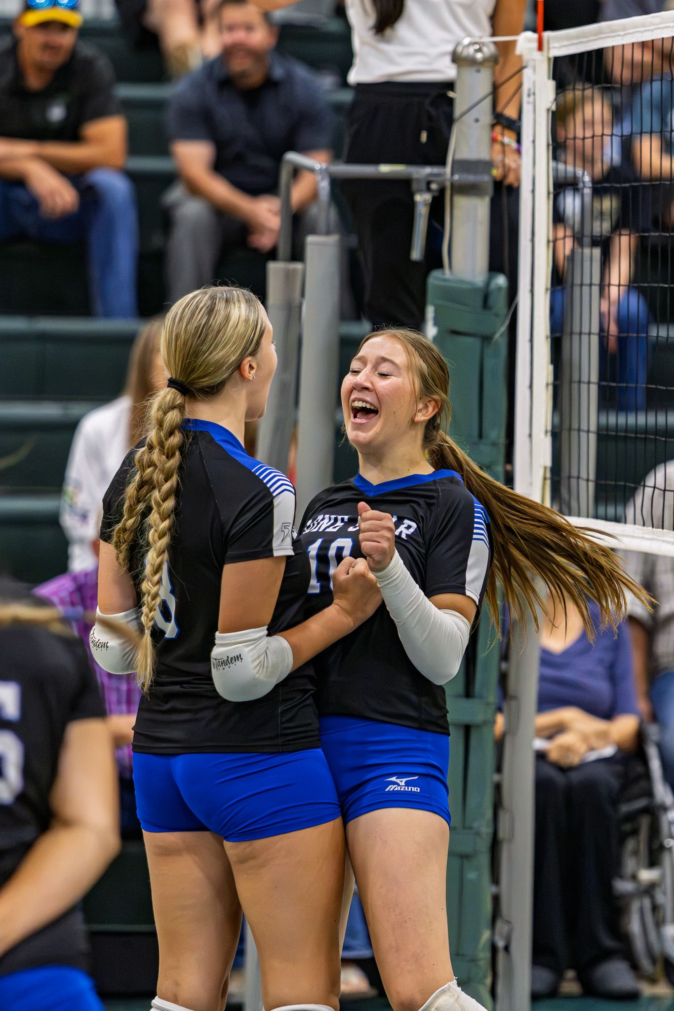 Two female volleyball players in black and blue Mizuno uniforms celebrating near the net during a match.