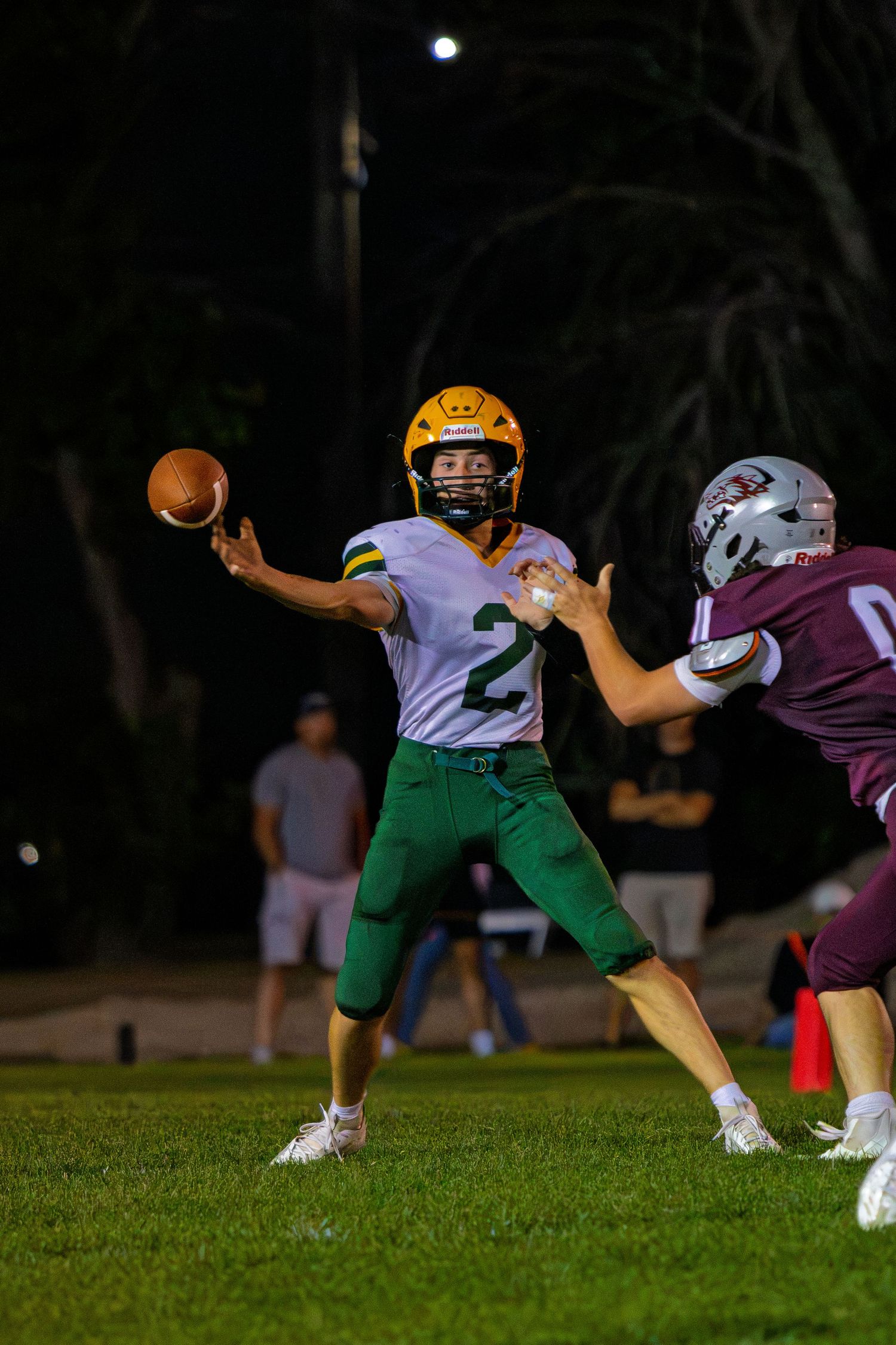 Youth football quarterback #2 in green and gold uniform throws a pass under pressure during a night game.