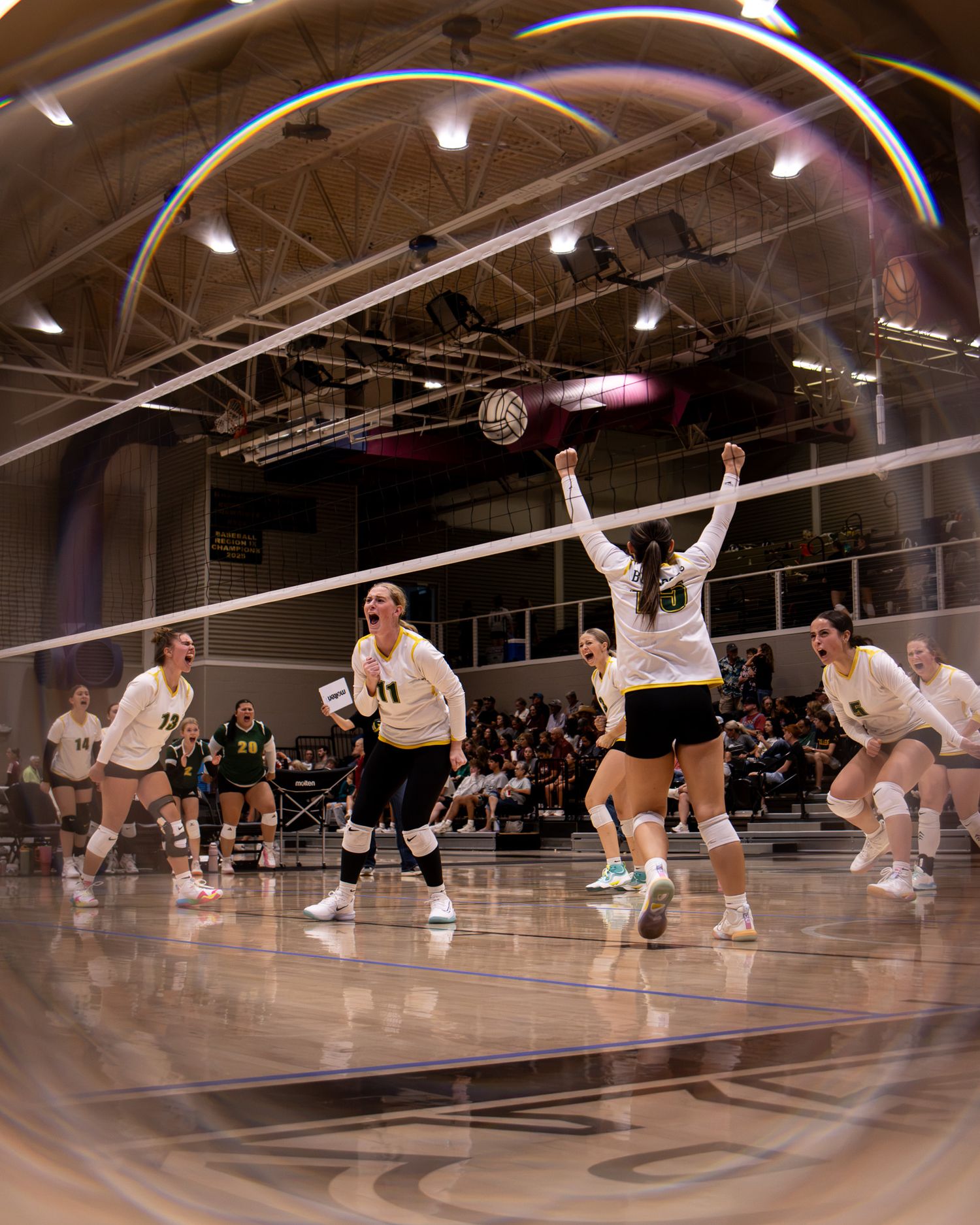 Women's volleyball team celebrates a point during an indoor match, with players raising arms near the net.