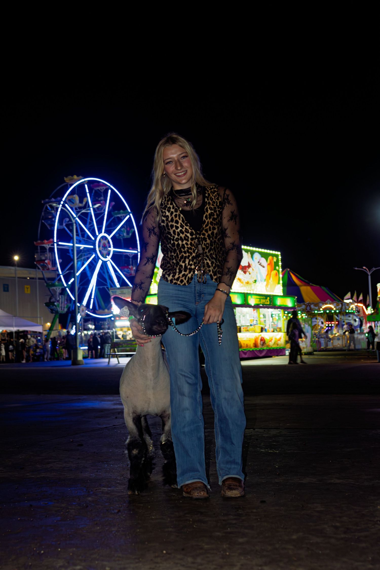 Blonde girl in leopard print top standing with a sheep at a night county fair with Ferris wheel.