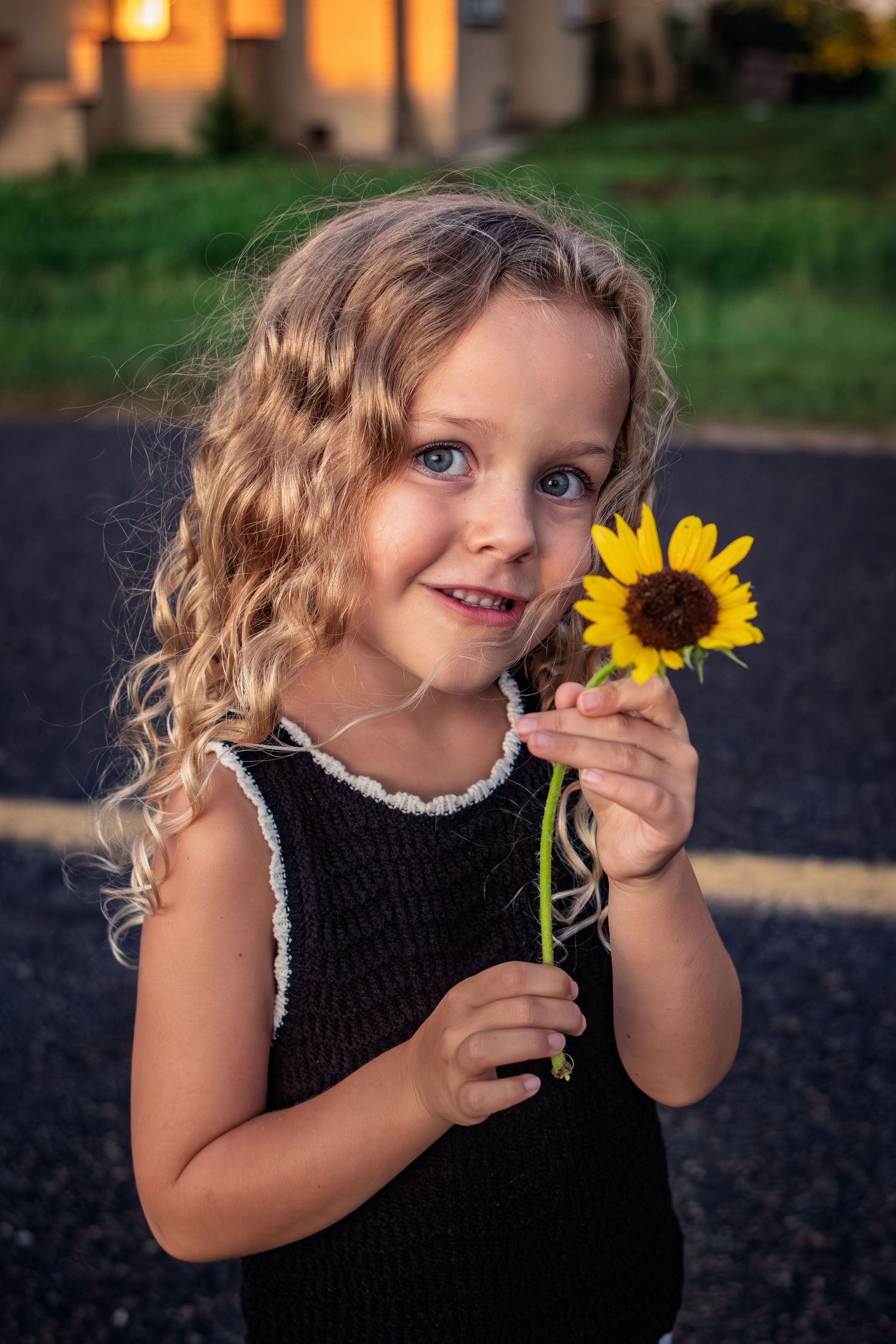 Adorable curly-haired blonde girl in black dress holding a sunflower at sunset outdoors.