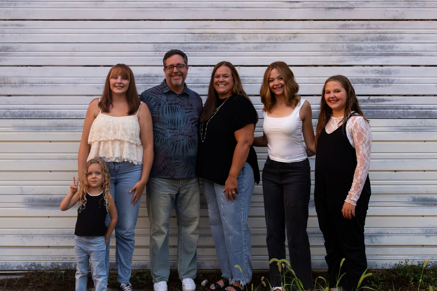 Family of six poses smiling in front of a rustic white wooden wall outdoors.