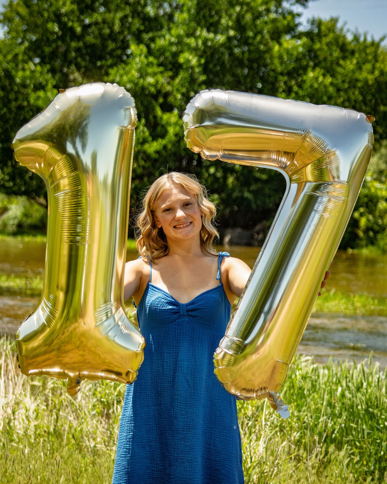 Smiling teenage girl in blue dress posing with gold number 17 balloons outdoors.