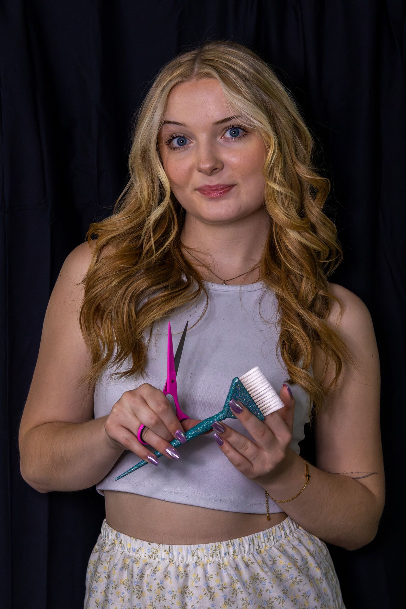 Blonde woman with curly hair holding pink scissors and comb for cosmetology portrait on dark background.