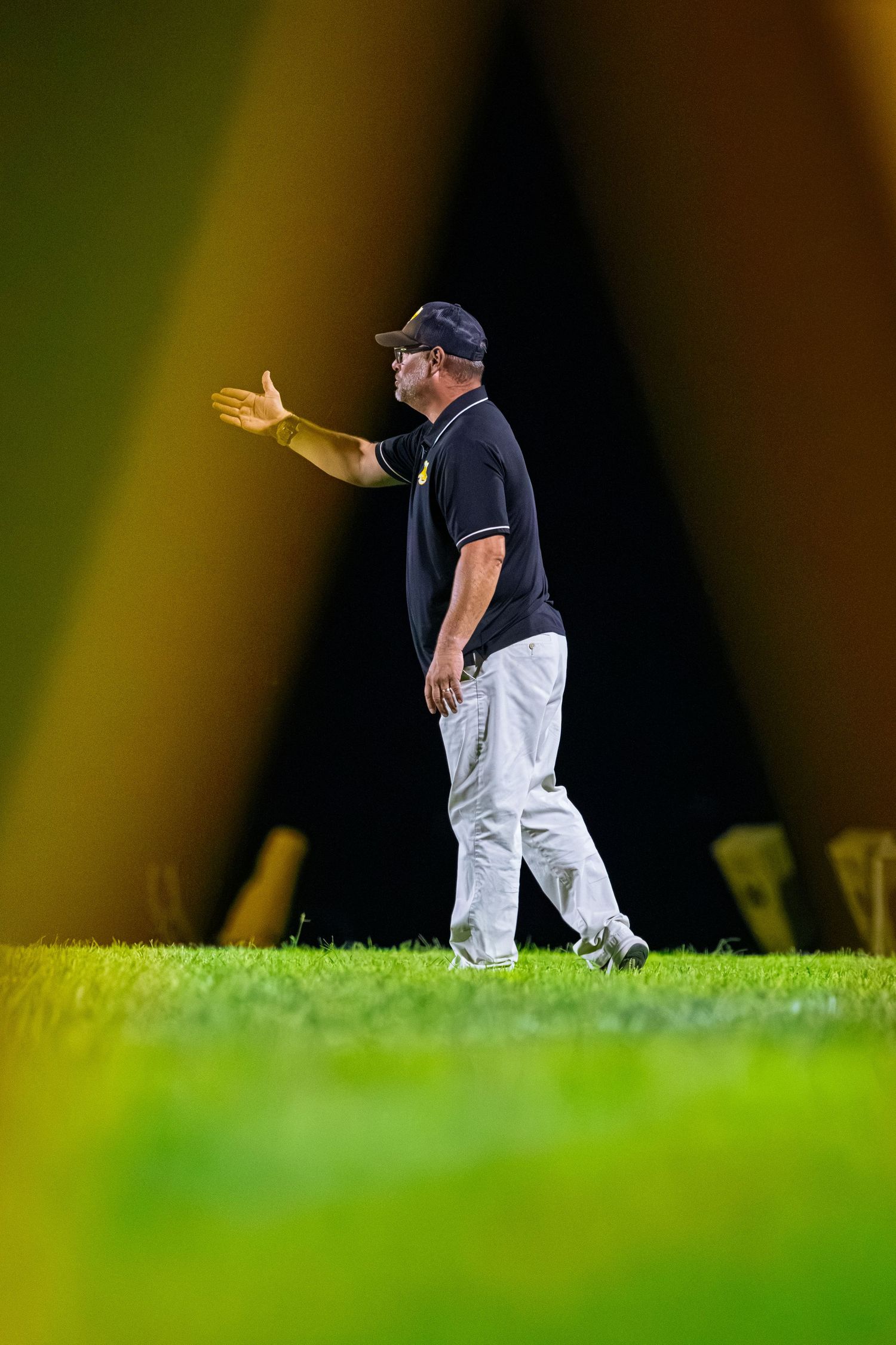 Football coach in black polo and white pants gesturing on the sideline at a night game.