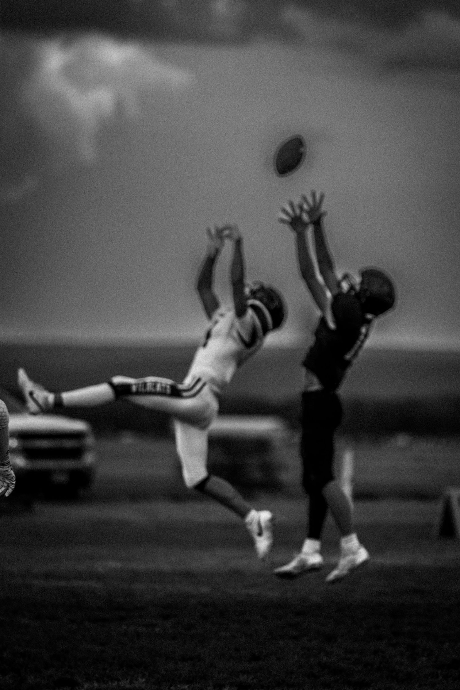 Two football players leap to contest a pass in a dramatic black and white action shot under stormy skies.