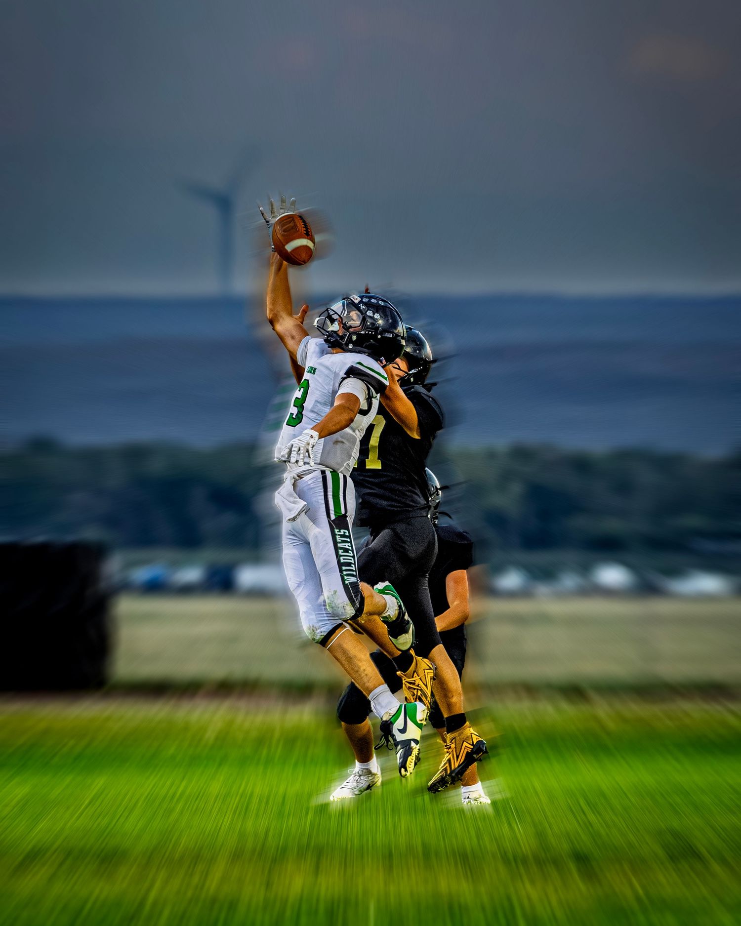 Football player in green uniform leaps to catch ball while defender attempts to block under dramatic sky.