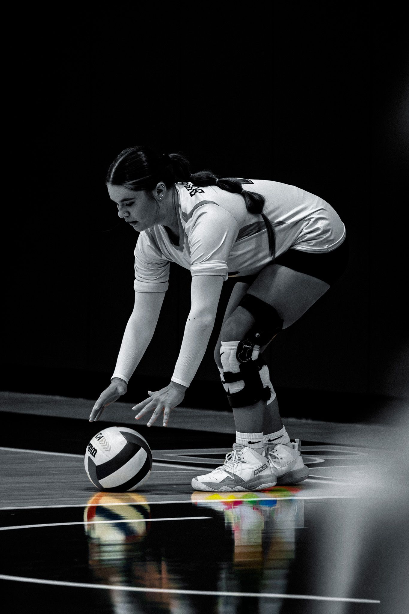 Female volleyball player in black and white photo bending down to dig a volleyball on a reflective court.