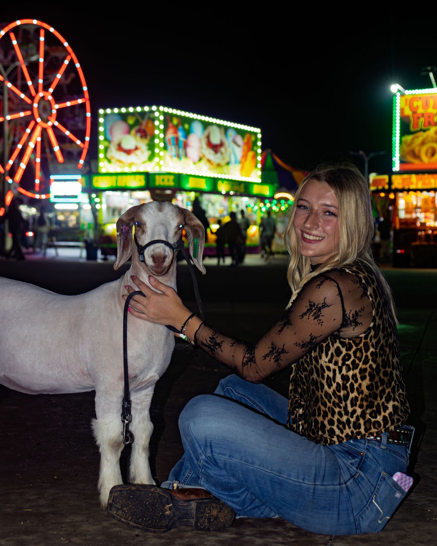 Blonde girl in leopard print top kneeling with a white goat at a colorful night county fair.