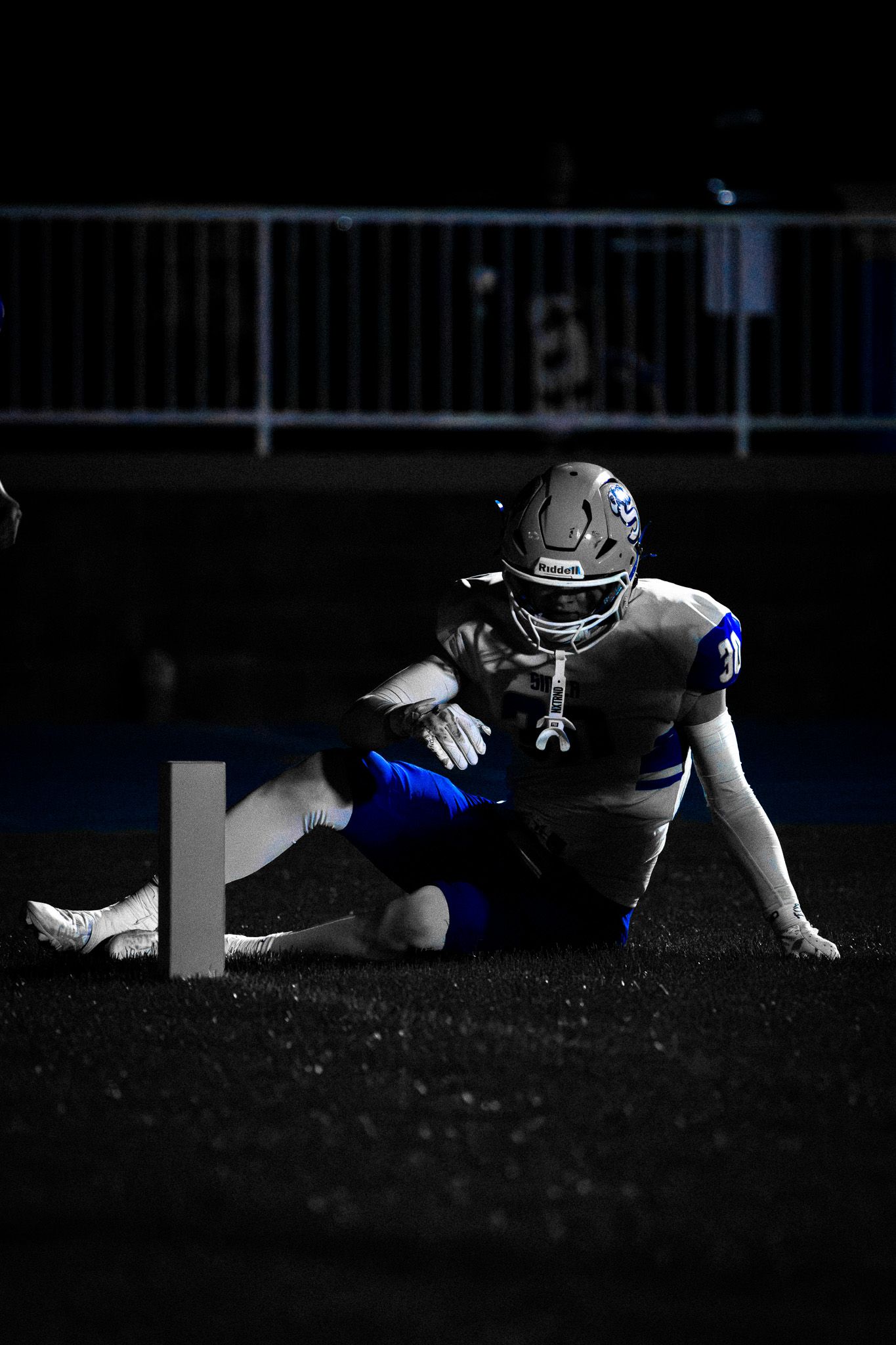 Football player in blue uniform crouches near yard marker in dramatic low-light night game photo.