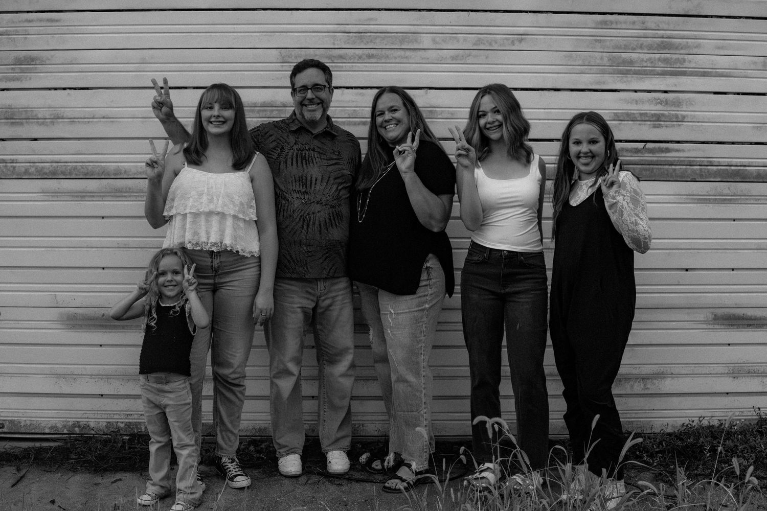 Black and white photo of a family of six making peace signs against a white wooden wall.