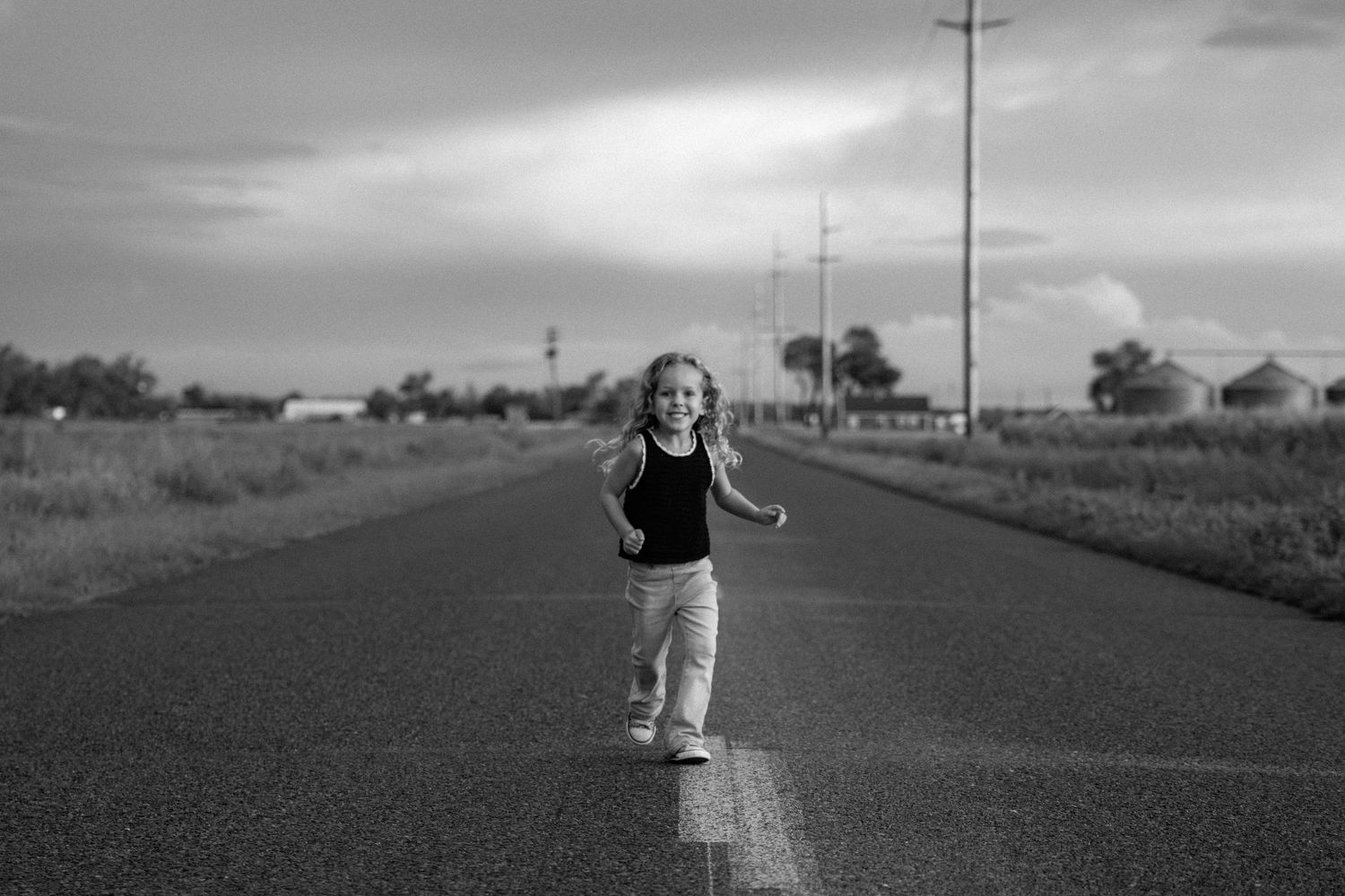Black and white photo of young curly-haired girl running on empty rural road under cloudy sky.