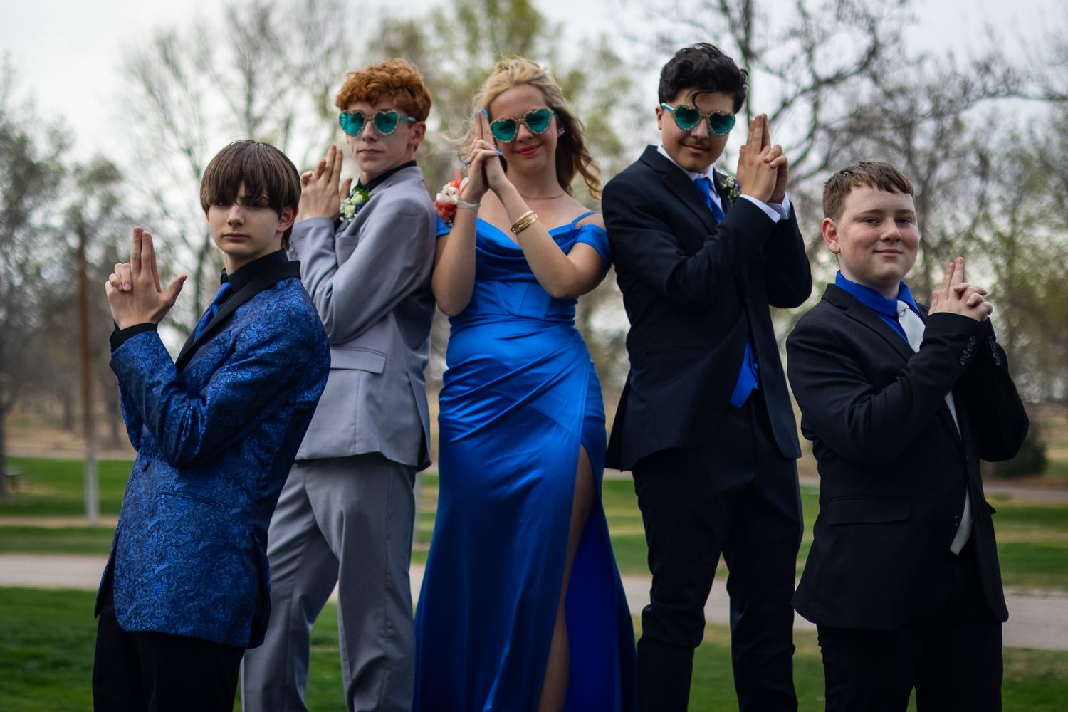 Five teenagers in formal prom attire pose playfully outdoors wearing blue sunglasses in a park setting.