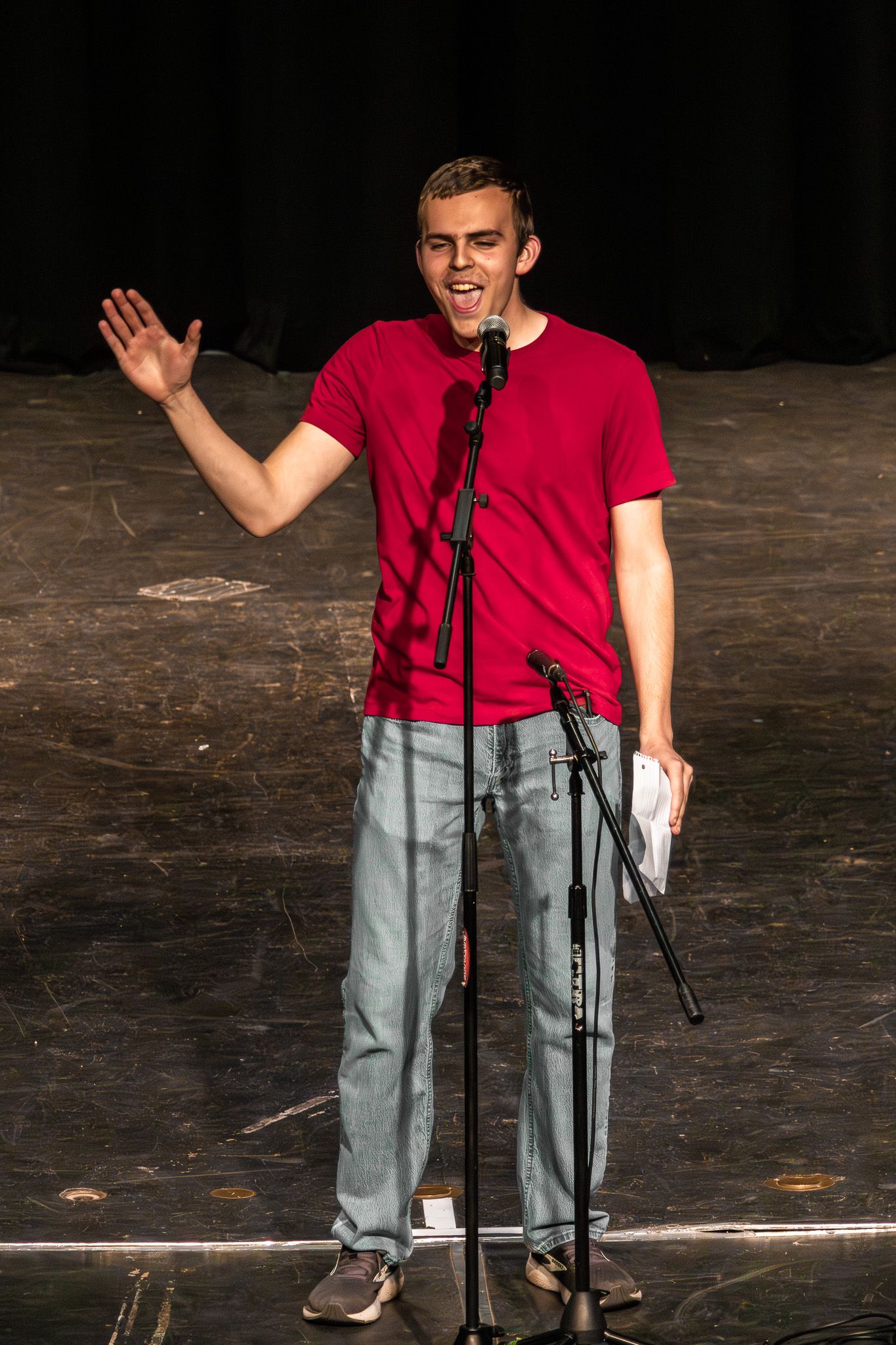 Young male performer in red t-shirt gesturing expressively at microphone stand on stage.