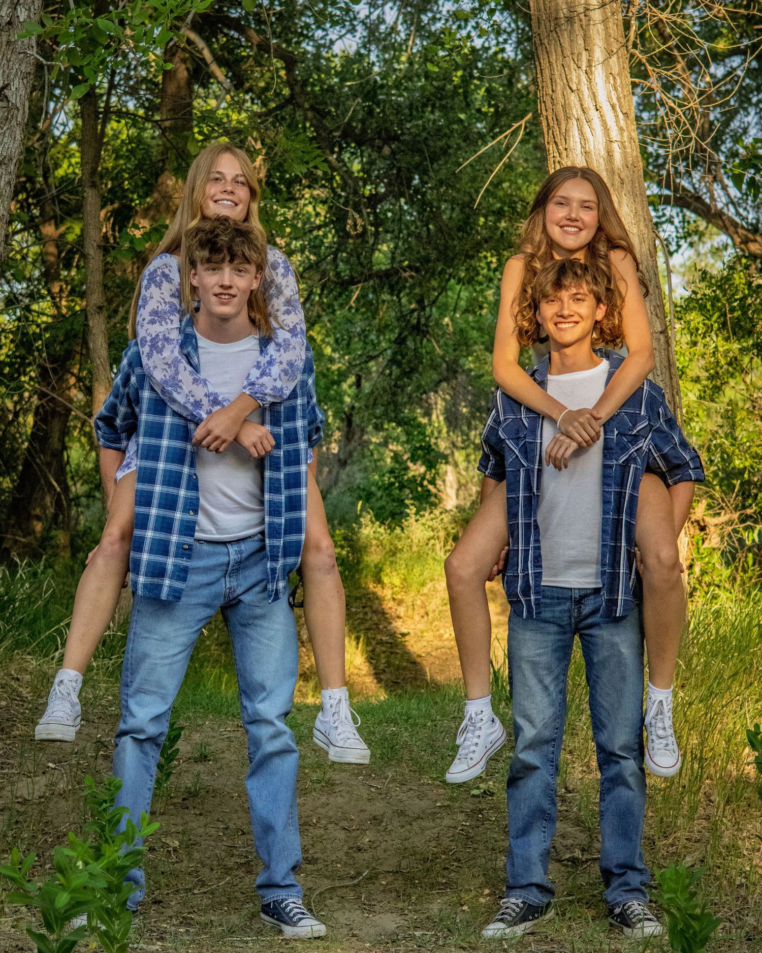 Two teenage couples laugh outdoors as boys give girls piggyback rides in a lush green forest setting.