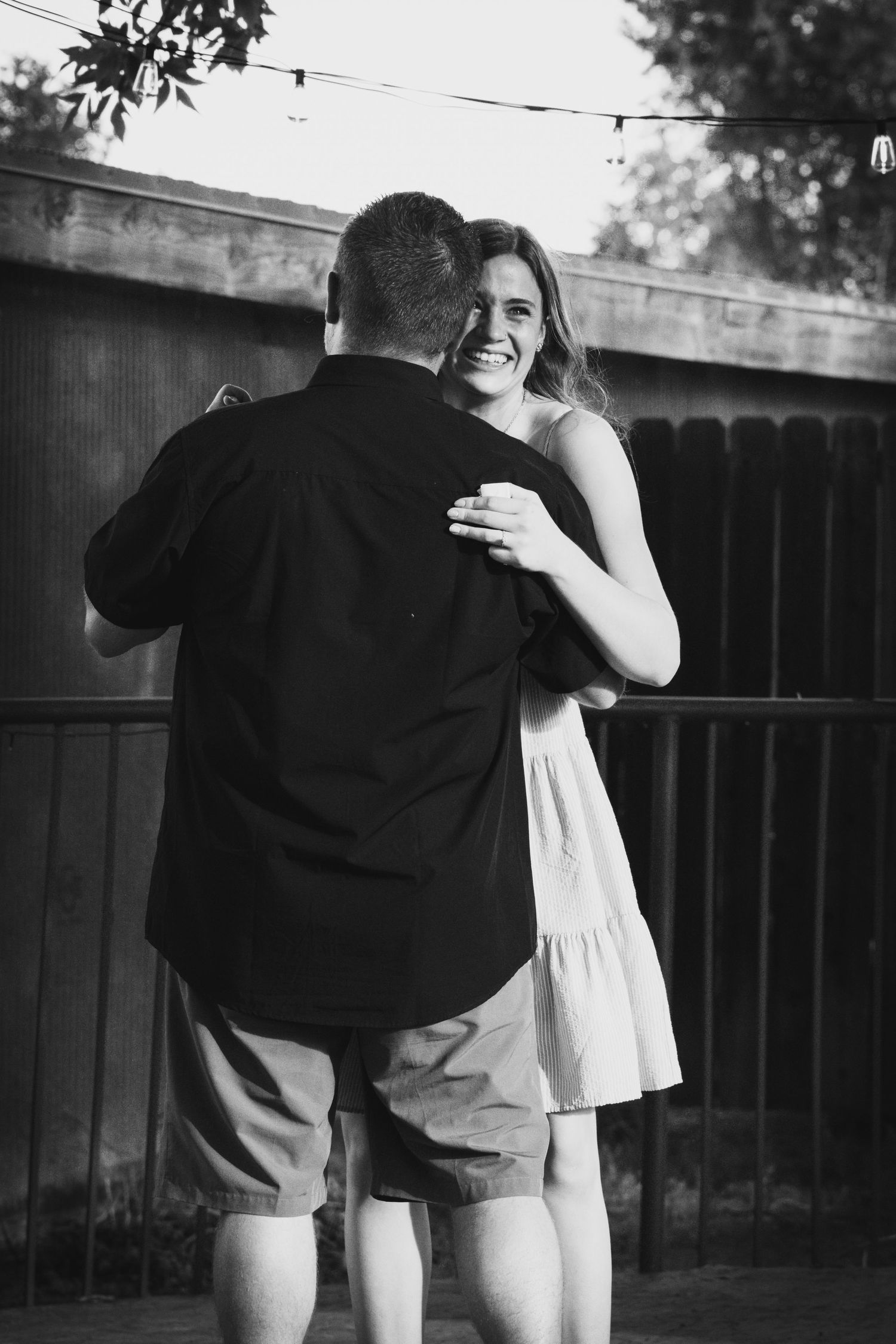 Black and white photo of smiling woman embracing man in backyard with string lights.