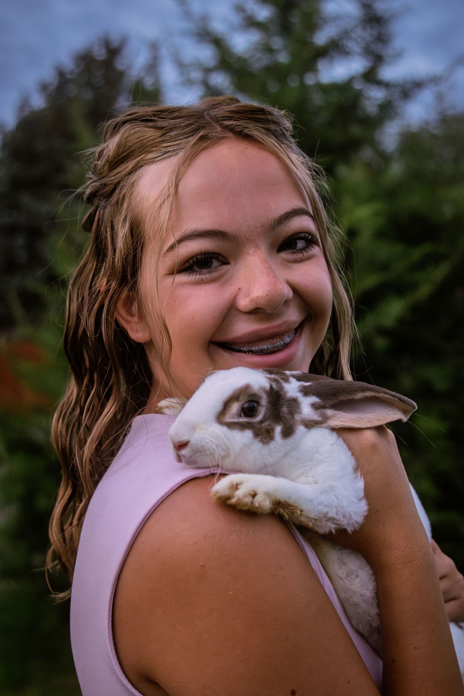 Smiling young girl with braces holding a white and brown rabbit close to her shoulder outdoors.