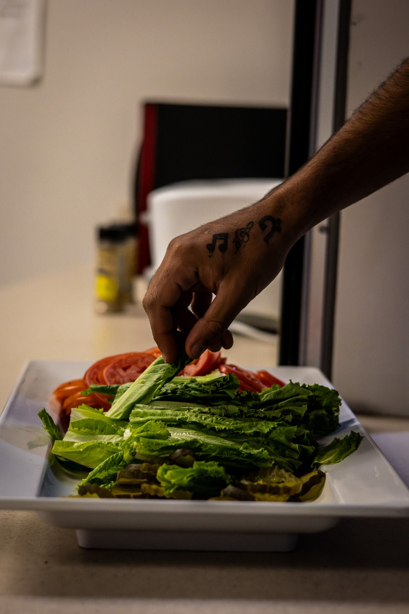 A tattooed hand arranges fresh romaine lettuce and tomatoes on a white rectangular plate.