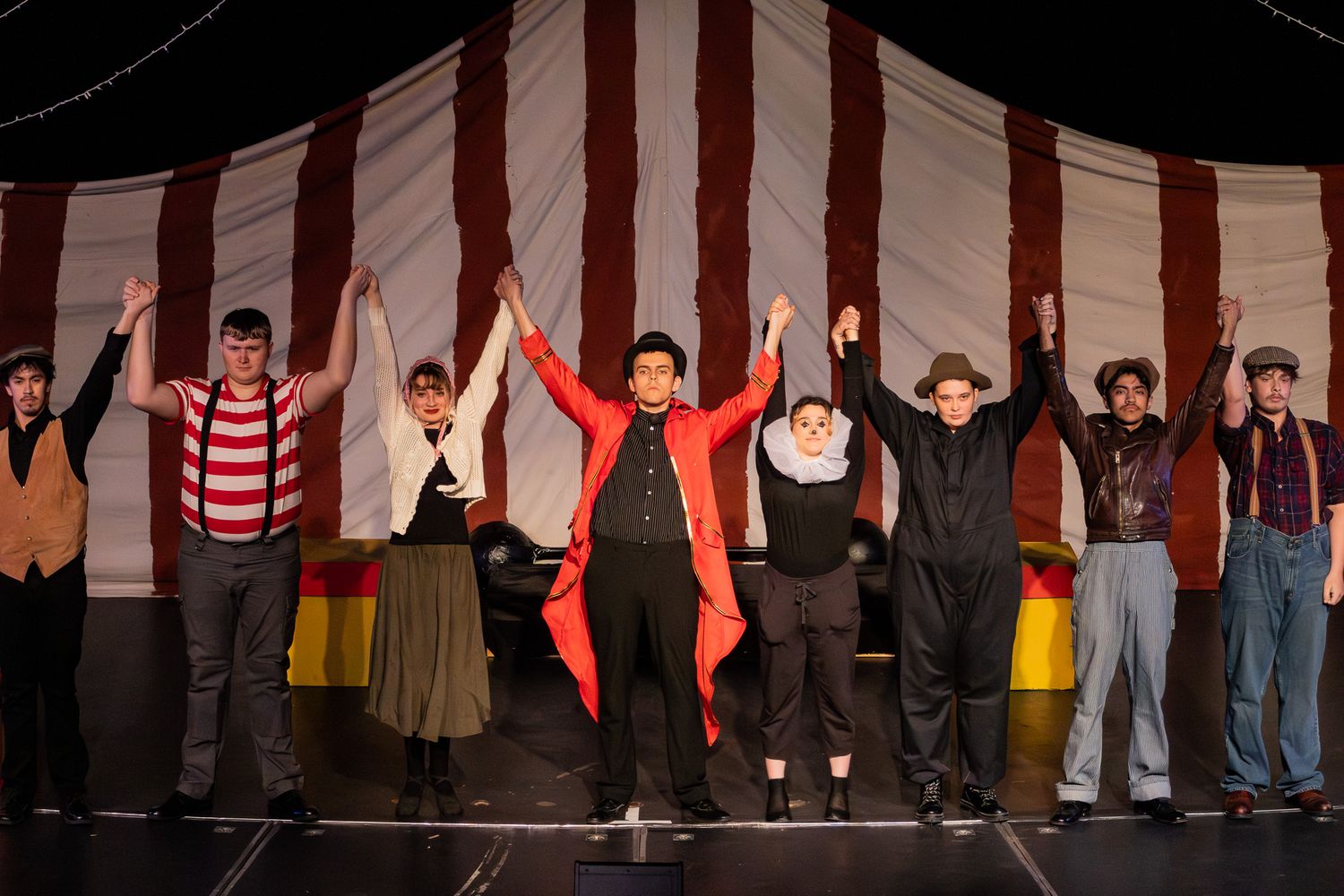 Theater cast takes a bow holding hands on stage in front of a striped circus tent backdrop.