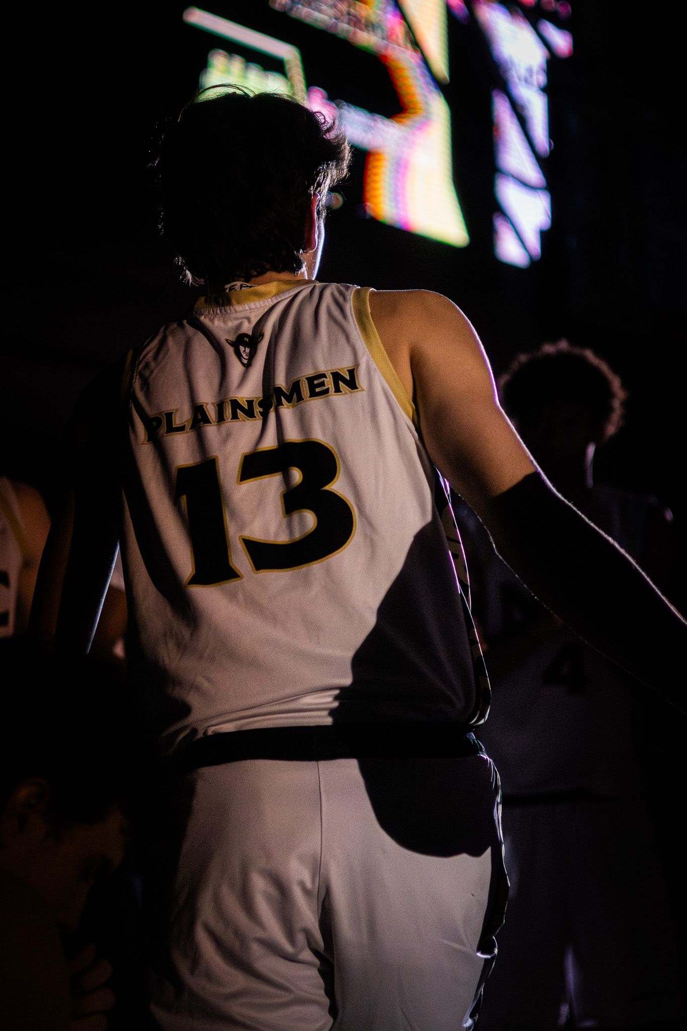 NJC Plainsmen player #13 walks in dark arena wearing white and gold jersey before a basketball game.