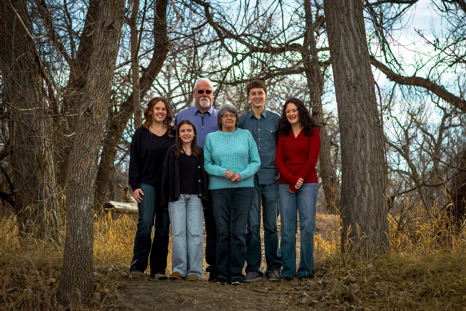 A family of six smiles together among bare trees in a natural outdoor winter setting.