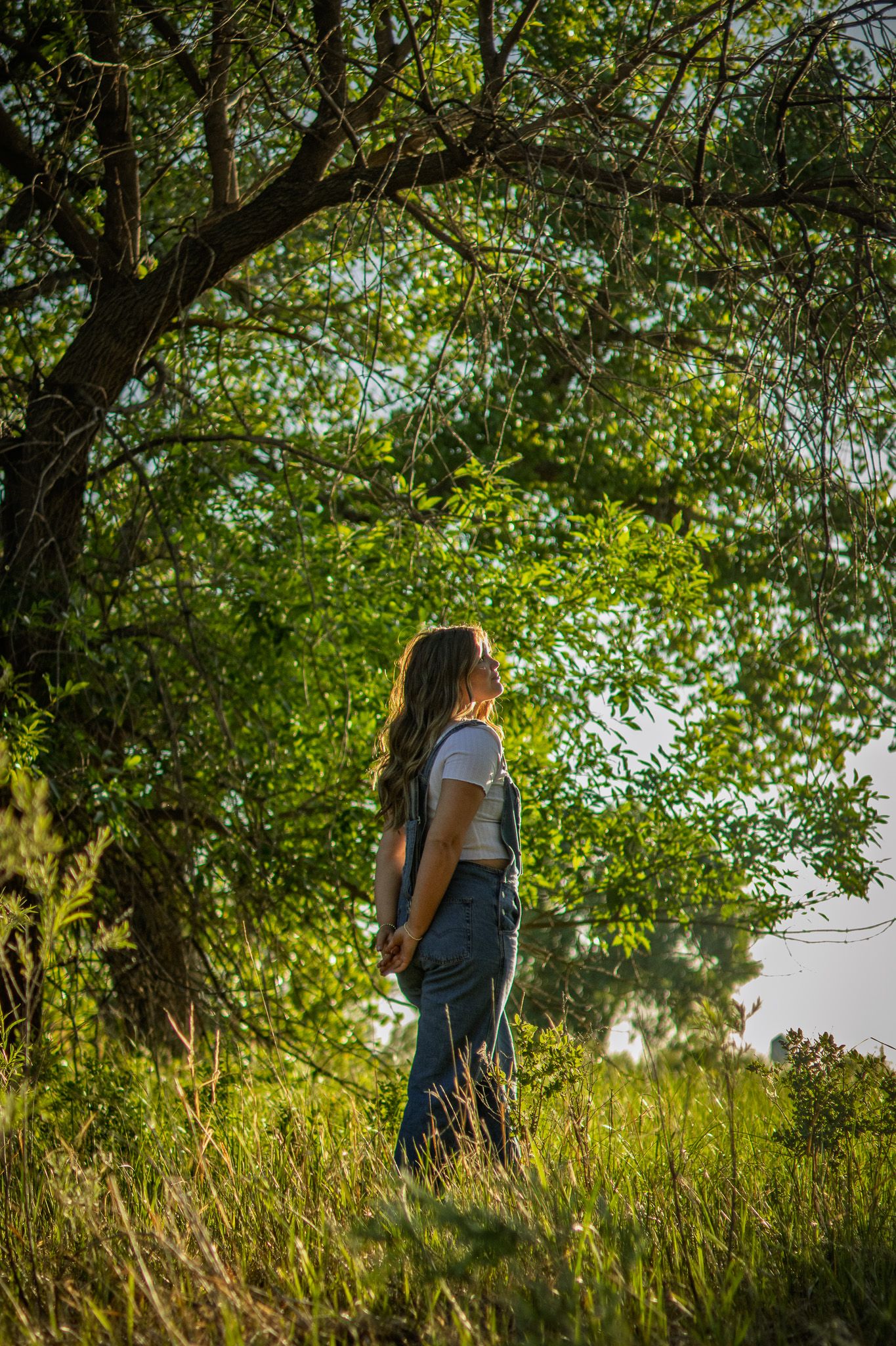 Young woman in white top and overalls standing in lush green field beneath sunlit tree canopy.