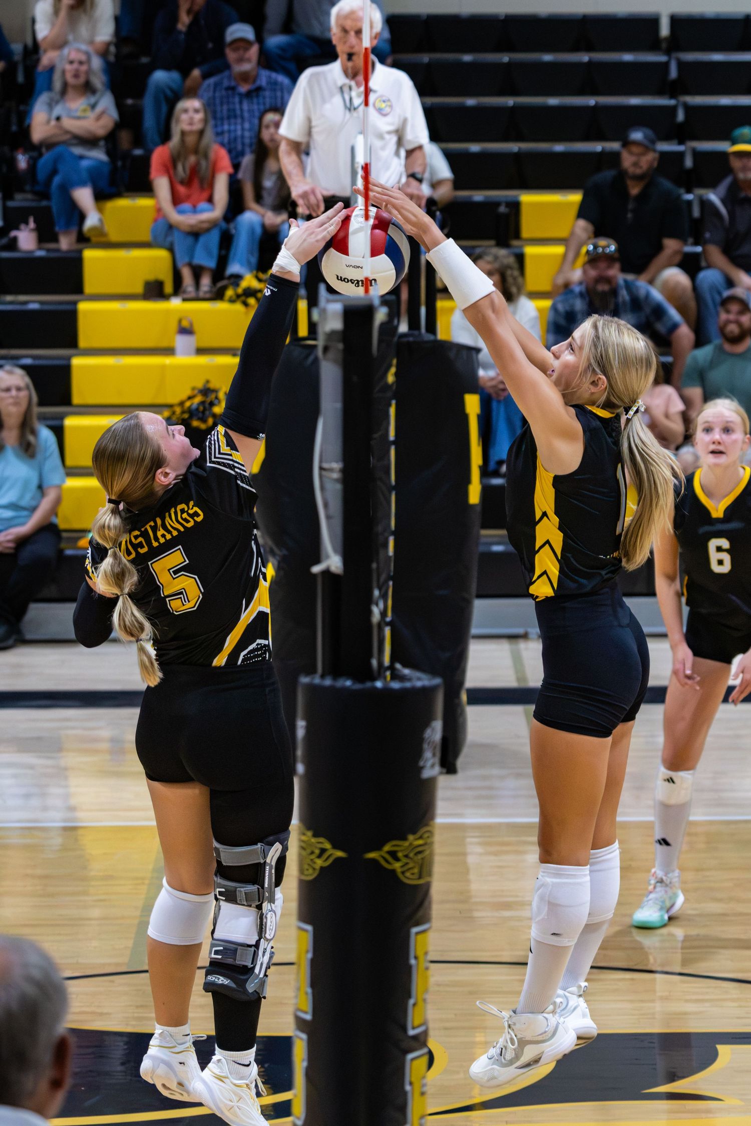 Two volleyball players in black and gold uniforms compete at the net during a high school game.