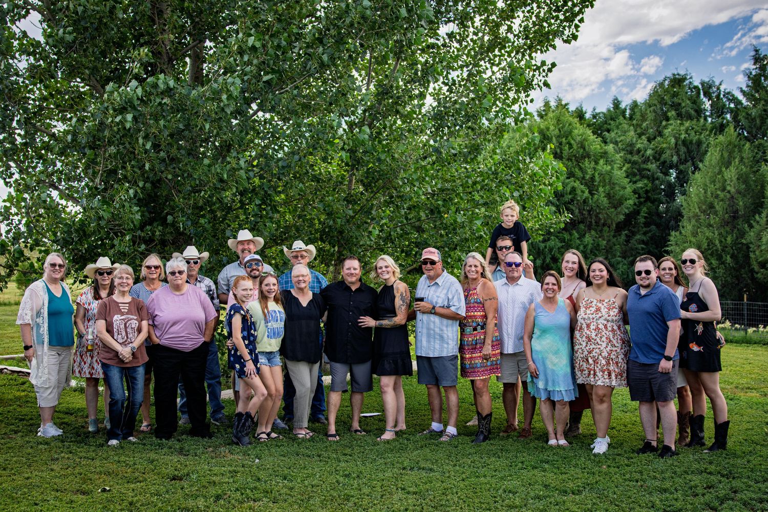 Large group of family and friends posing together on green lawn surrounded by tall trees.