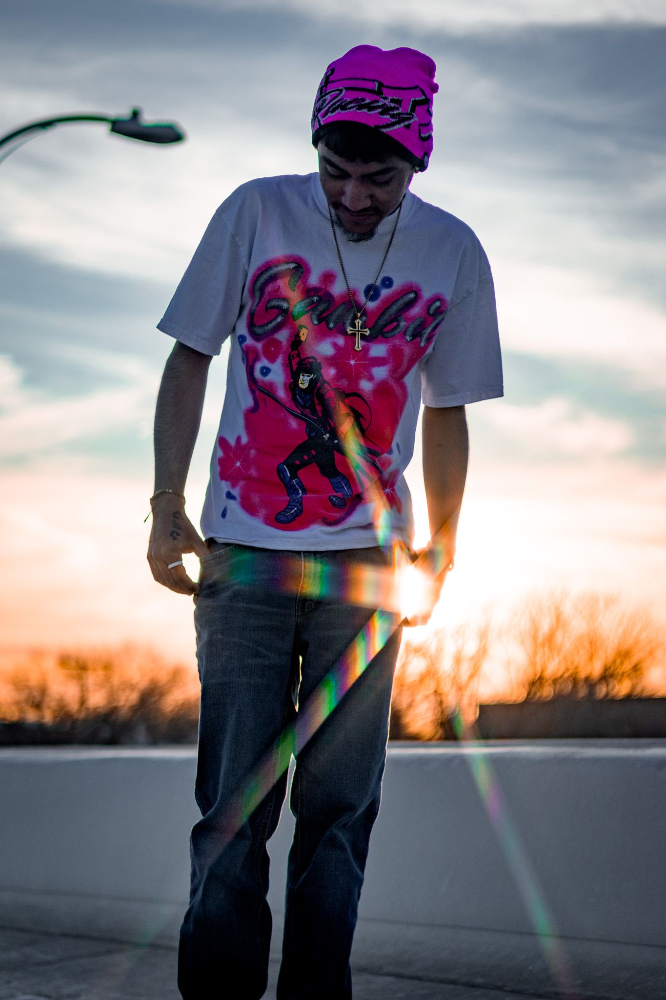 A man in a pink beanie and graphic tee stands outdoors with colorful lens flare at golden hour.