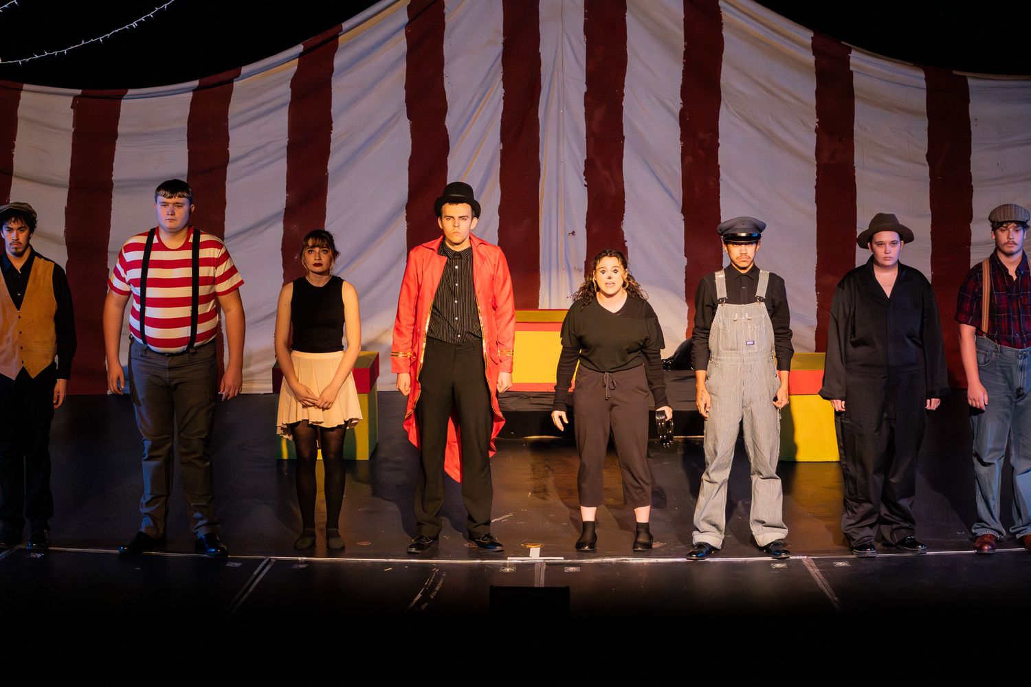 Theater cast performs together on stage in front of a red and white striped circus tent backdrop.