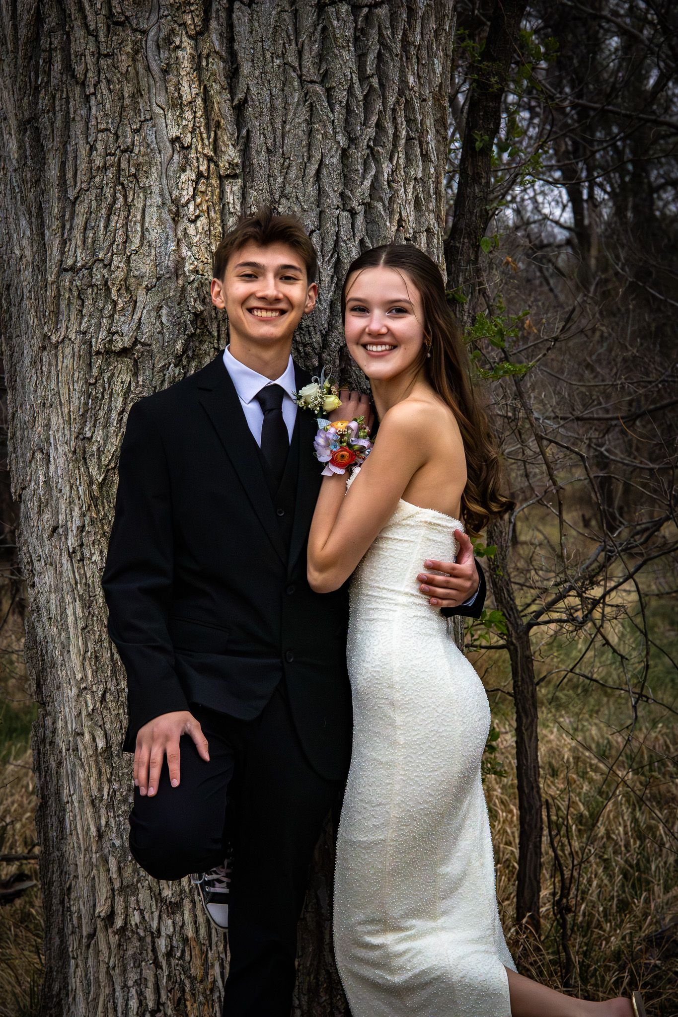 A young couple dressed in formal prom attire pose together smiling in front of a large tree.