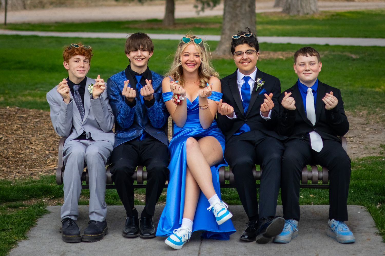 Five smiling teenagers in prom attire sit together on a park bench making heart gestures with their hands.