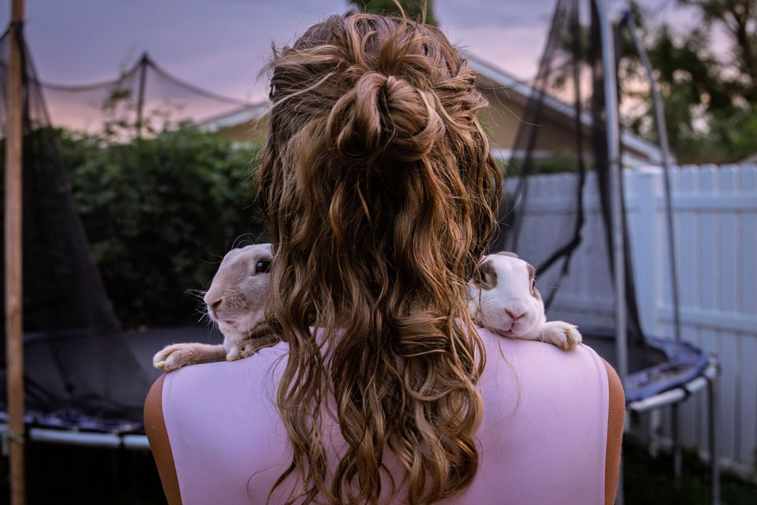 Girl with wavy hair has two white rabbits perched on her shoulders in a backyard at dusk.