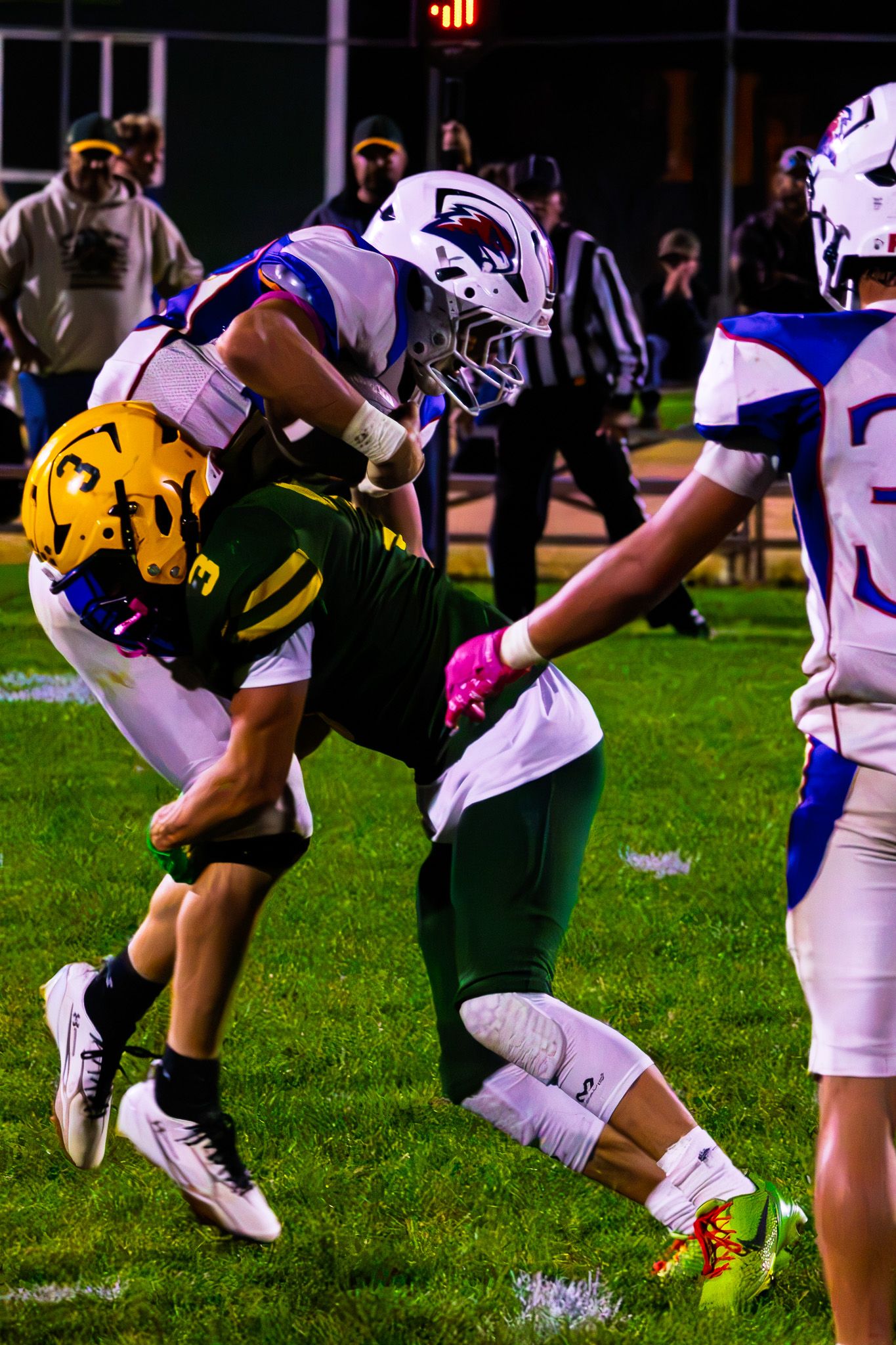 White-uniformed football player tackles green and gold opponent during an intense night game.