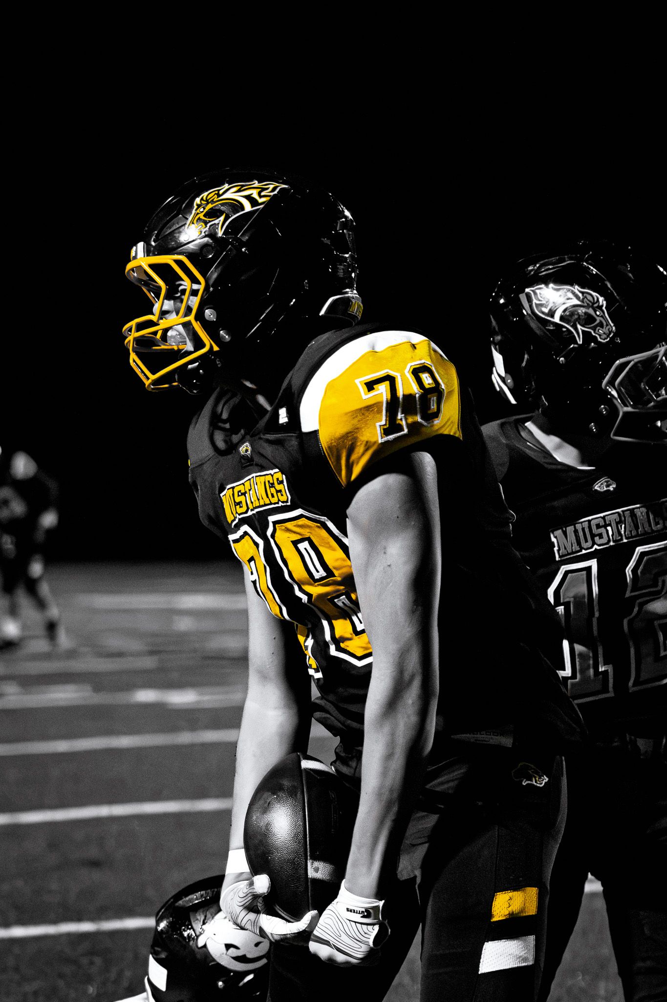 Two Mustangs football players in black and gold uniforms stand back to back on field at night.