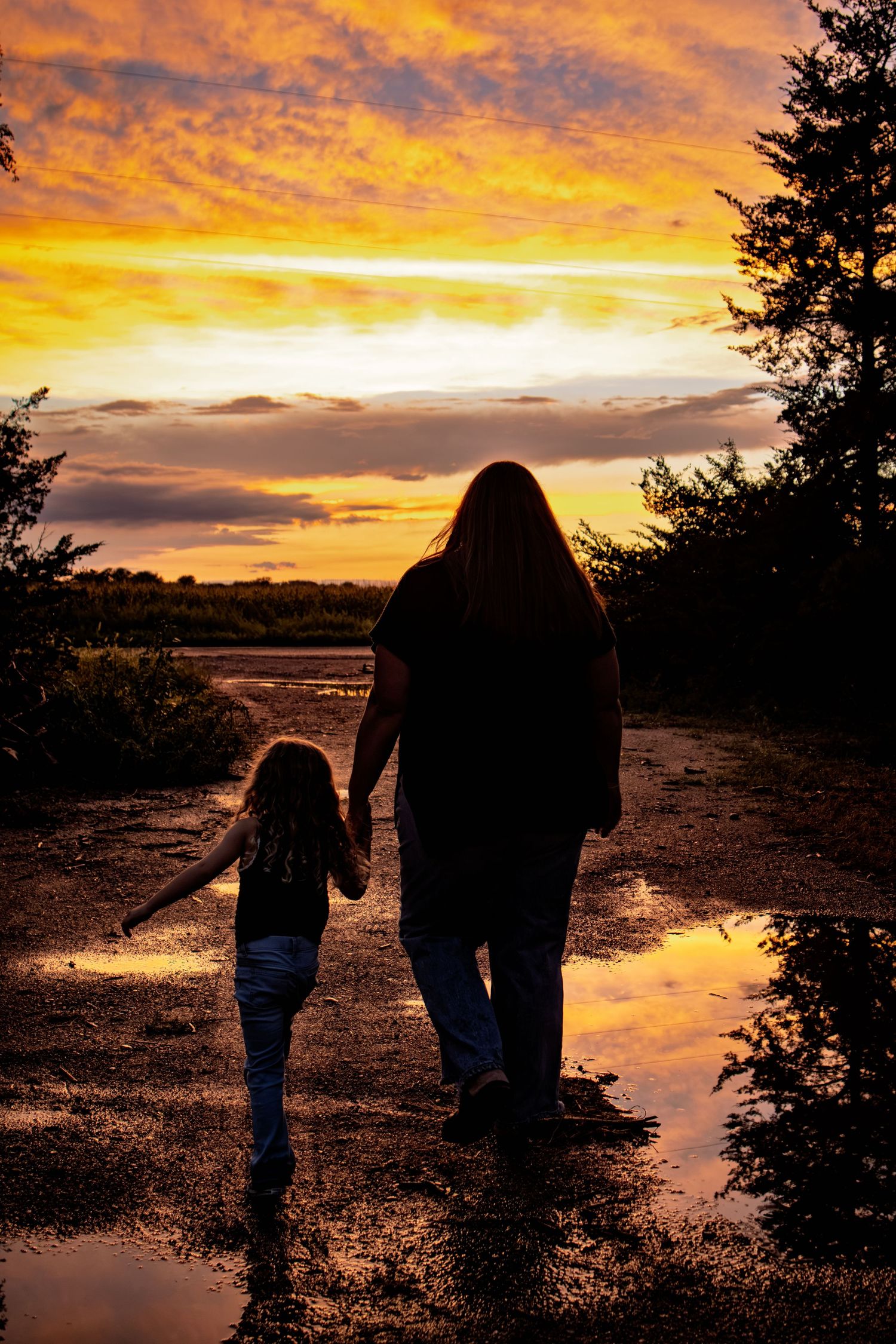 Silhouette of mother and daughter holding hands walking on wet path at vibrant golden sunset.
