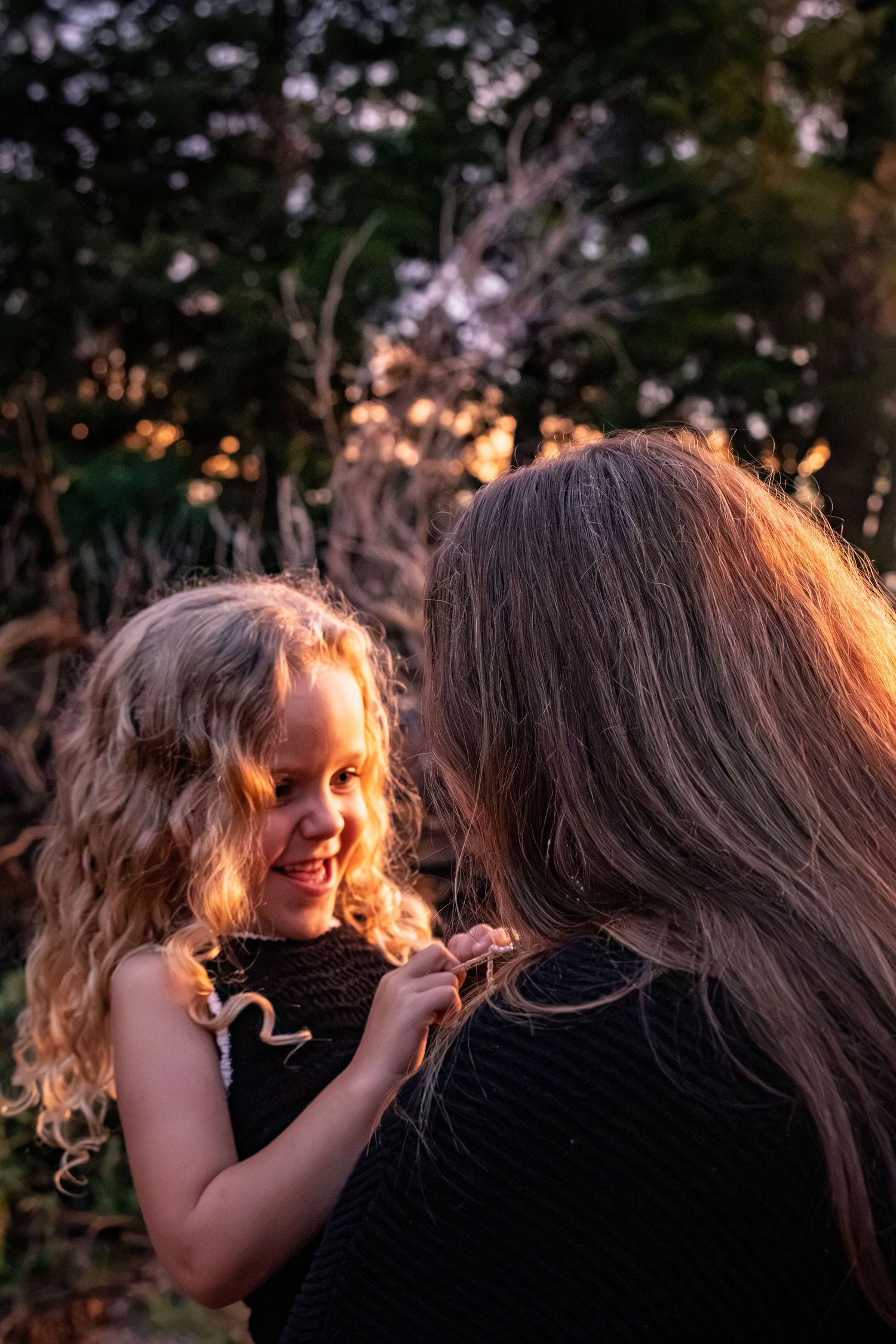Young curly-haired girl smiling at her mother in warm golden sunset light outdoors.