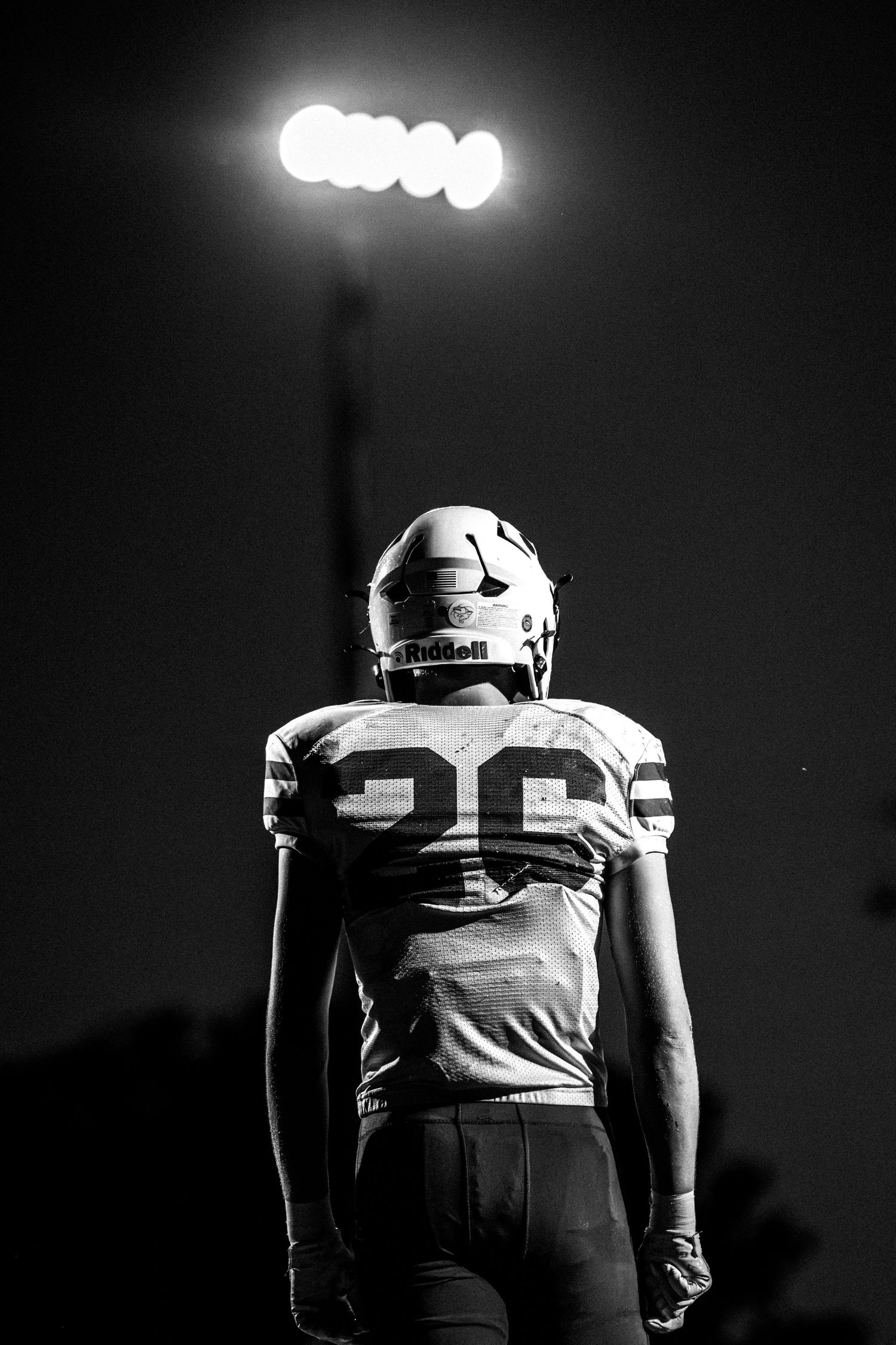 Black and white dramatic photo of football player #26 in Riddell helmet standing under bright stadium lights at night.