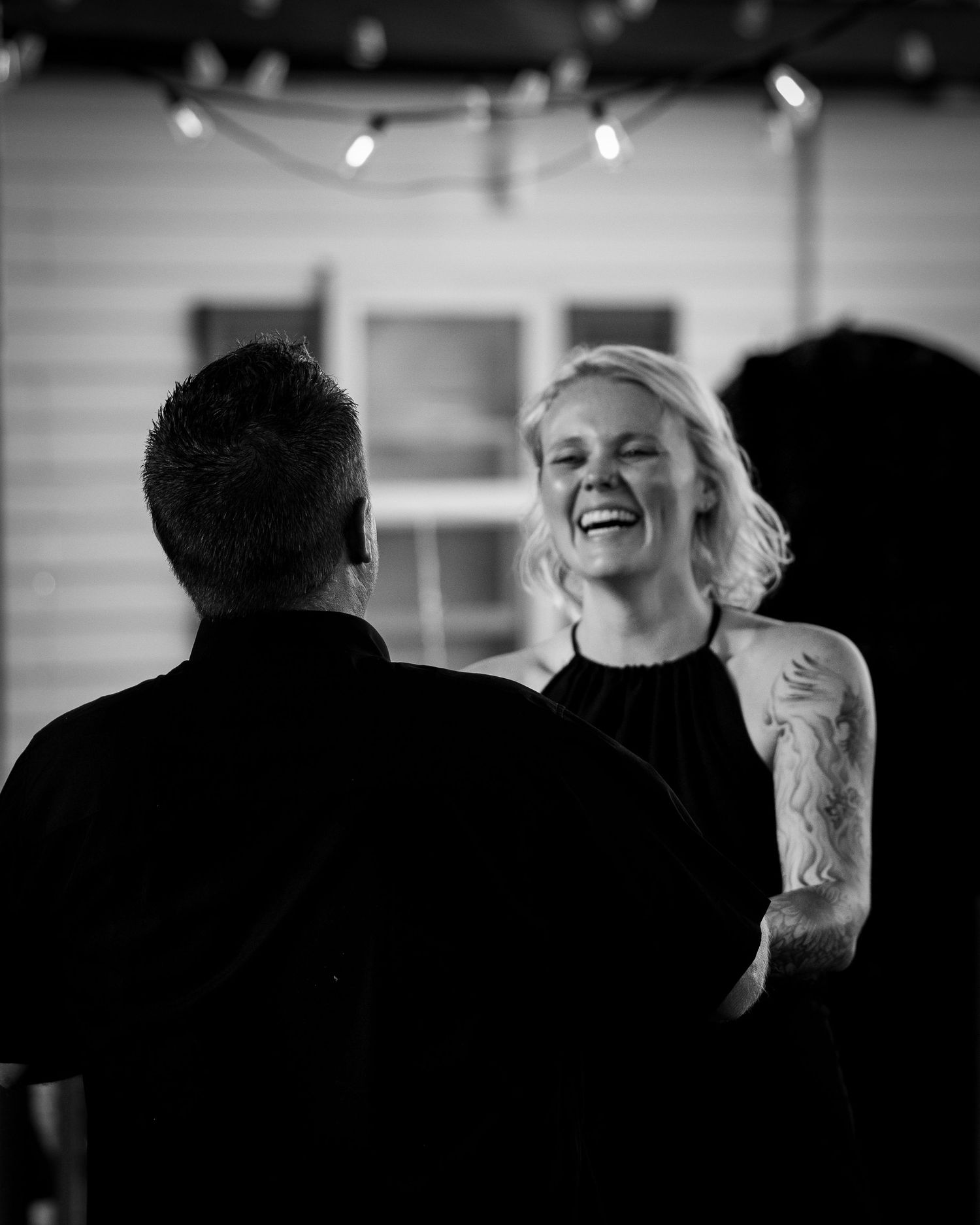 Black and white photo of a laughing blonde woman with tattoos dancing at an indoor event with string lights.