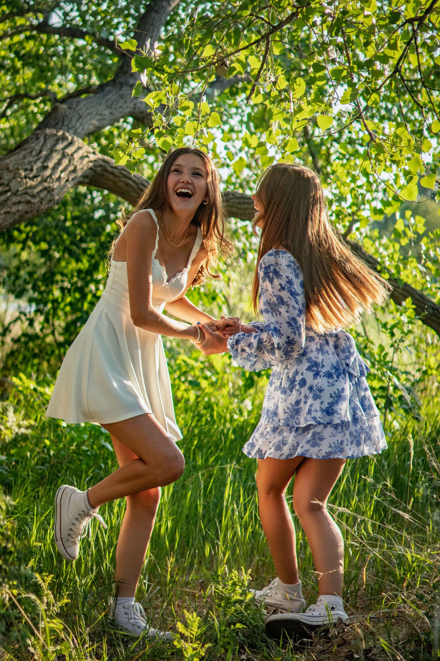 Two laughing girls in summer dresses hold hands and dance together under a sunlit green tree outdoors.