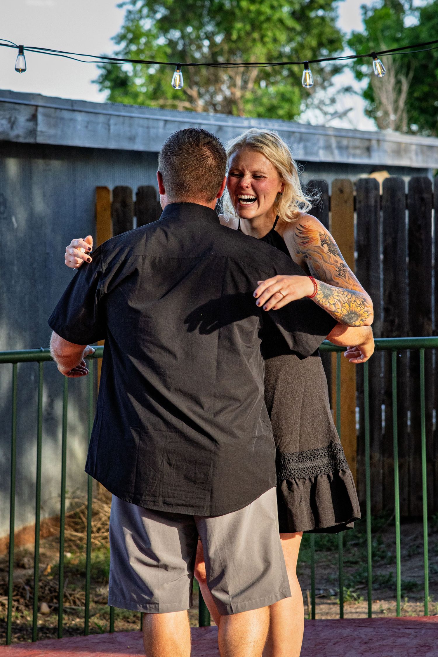 Laughing tattooed blonde woman dancing with man outdoors under string lights at dusk.