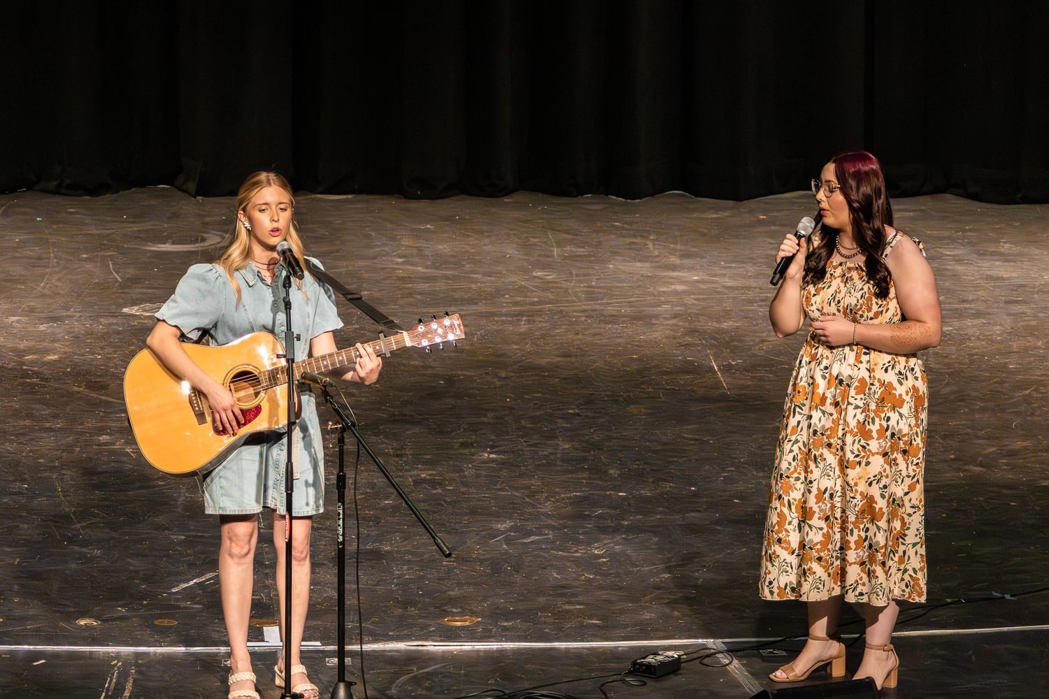 Two young women perform on stage, one playing acoustic guitar and one singing into a microphone.
