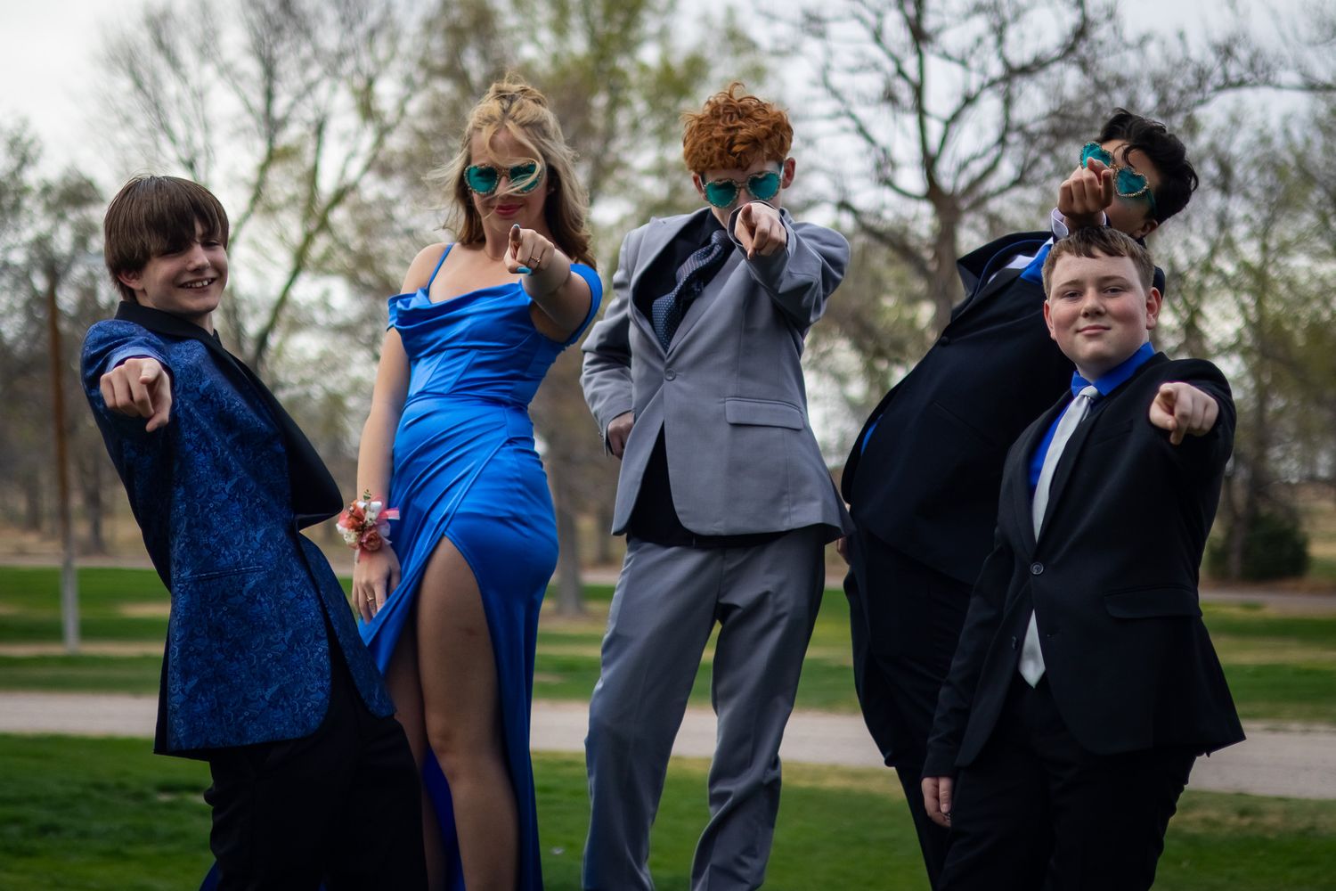 Group of five prom-dressed teens striking fun poses in a park, wearing stylish suits and a blue gown.