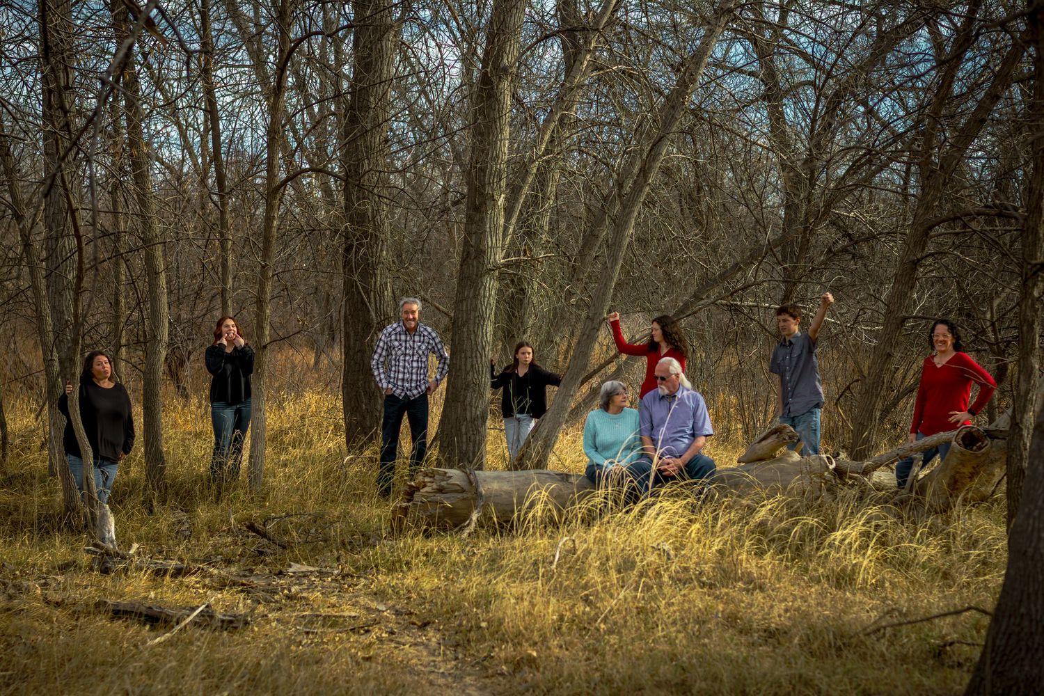 A large family group poses casually among bare trees and tall grass in an autumn woodland setting.