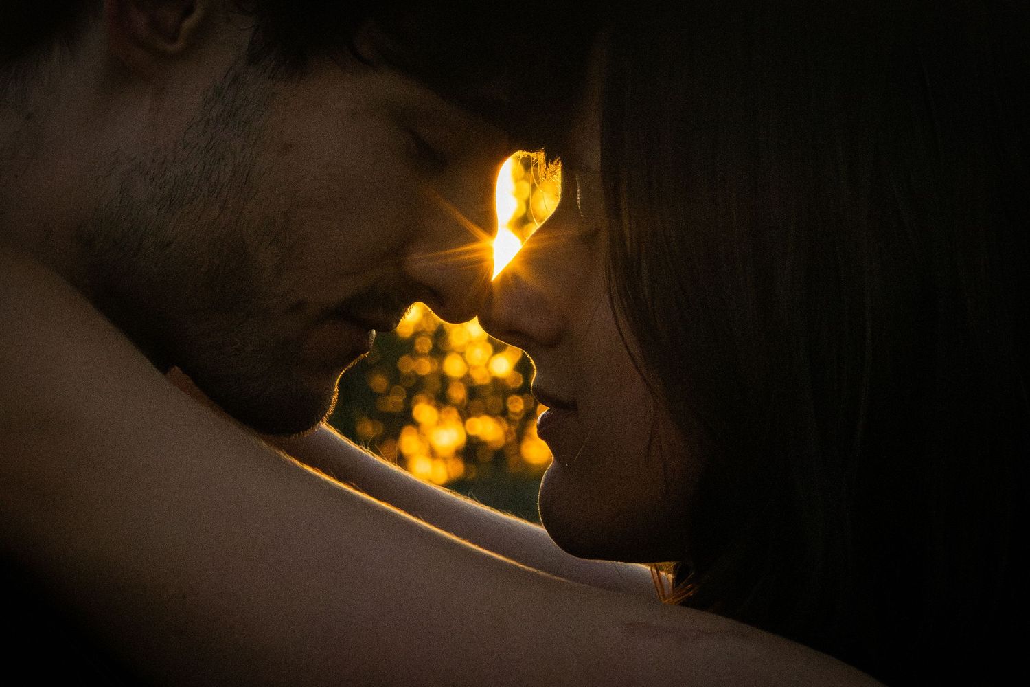 A couple shares an intimate moment in darkness with warm golden bokeh lights glowing between them.